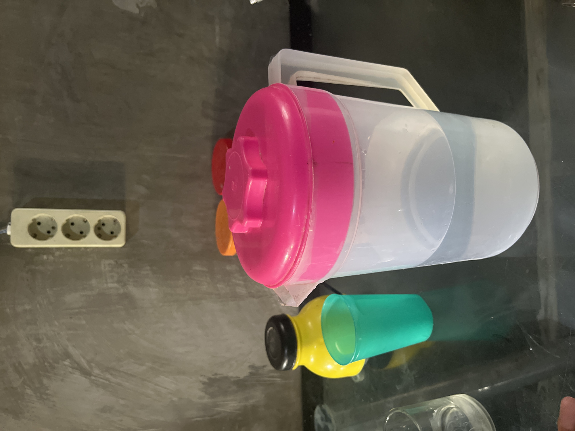 This image depicts a domestic setting, likely a kitchen or dining area, in Laban, Indonesia. In the foreground, a clear plastic pitcher with a bright pink lid contains a light blue liquid, presumably water. To the left of the pitcher, a vibrant green plastic cup stands empty. Behind these items, a bright yellow bottle with a black lid is visible. In the background, three brightly colored lids (orange and red) are stacked. The wall behind the objects is textured and gray, with a white electrical outlet featuring three sockets mounted on it. The surfaces appear to be dark and reflective, suggesting a countertop or table. The overall lighting is dim, creating shadows. There are no people visible in the frame.
