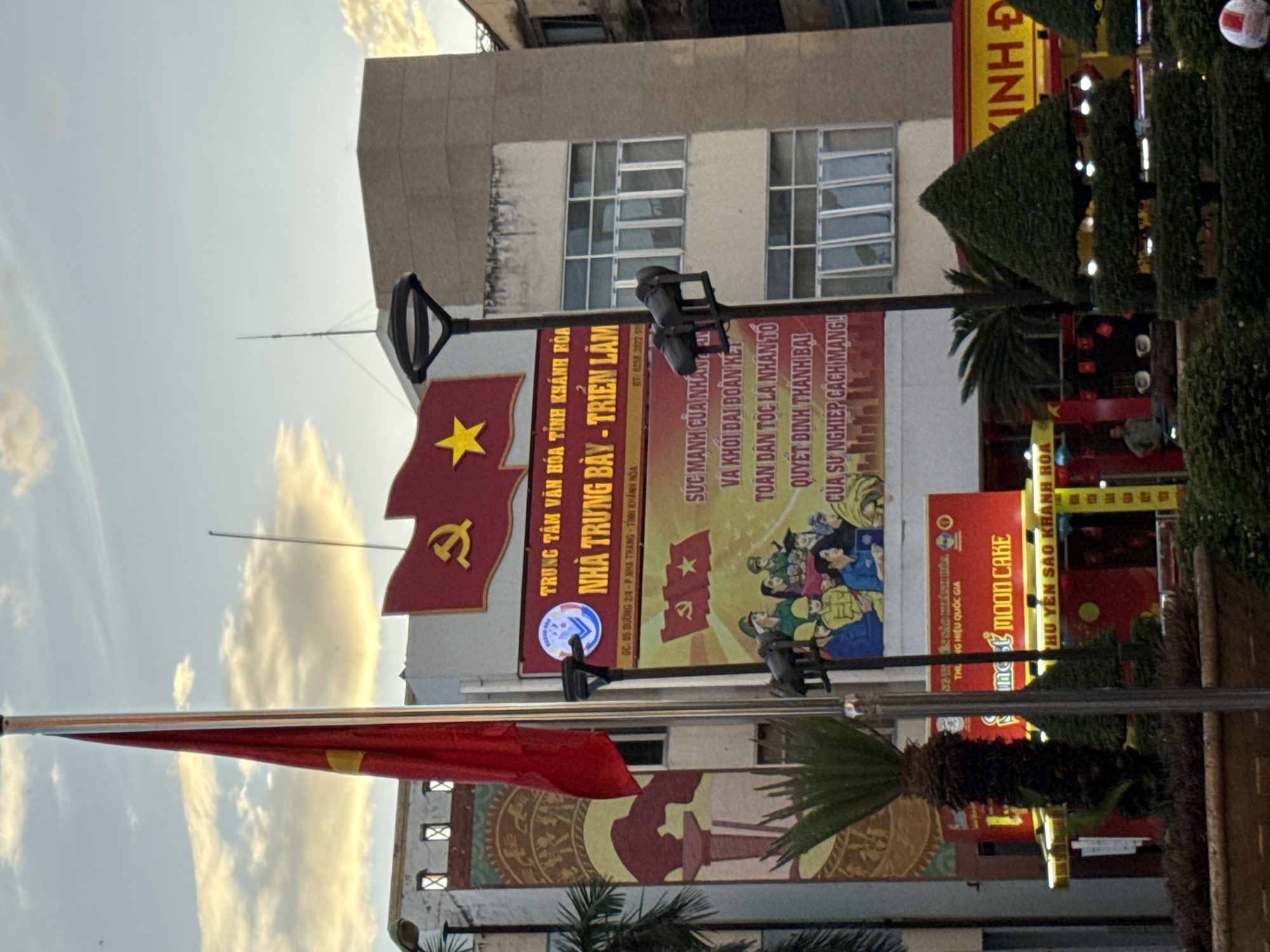 The image captures a street scene in Nha Trang, Vietnam, under a cloudy sky with golden hues suggesting either dawn or dusk. Dominating the foreground is a tall flagpole displaying a red flag with a yellow star, a prominent symbol of the Socialist Republic of Vietnam. To the right of the flag, a building displays a large banner with Vietnamese text and imagery consistent with socialist propaganda, featuring the hammer and sickle symbol and a depiction of people in military-style uniforms.
Below the main banner, a red sign with yellow lettering advertises "MOON CAKE" and further Vietnamese text. In front of this building, neatly trimmed evergreen topiaries and a potted palm tree contribute to the urban landscaping. A street lamp with two lights is visible in the mid-ground, positioned between the flagpole and the banner. The background shows multi-story buildings with windows, characteristic of urban architecture. The overall atmosphere suggests a public space, possibly a plaza or square, in Nha Trang.