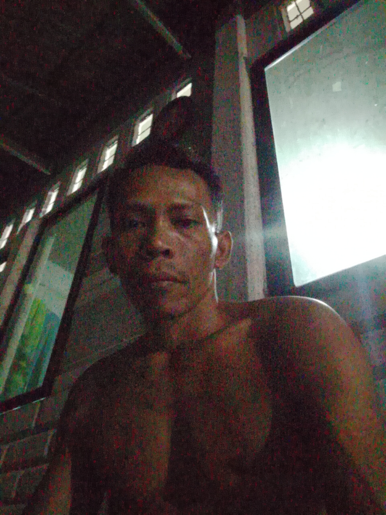 A photograph taken in Bukit Ketok, Indonesia depicts a shirtless man indoors. He has short dark hair and appears to be in his late 20s to early 30s. The image is taken at a low angle, suggesting the camera is below the subject. The background shows windows, one of which is letting in a substantial amount of light. There is minimal other discernible detail in the room. The overall image quality is grainy and somewhat dark.