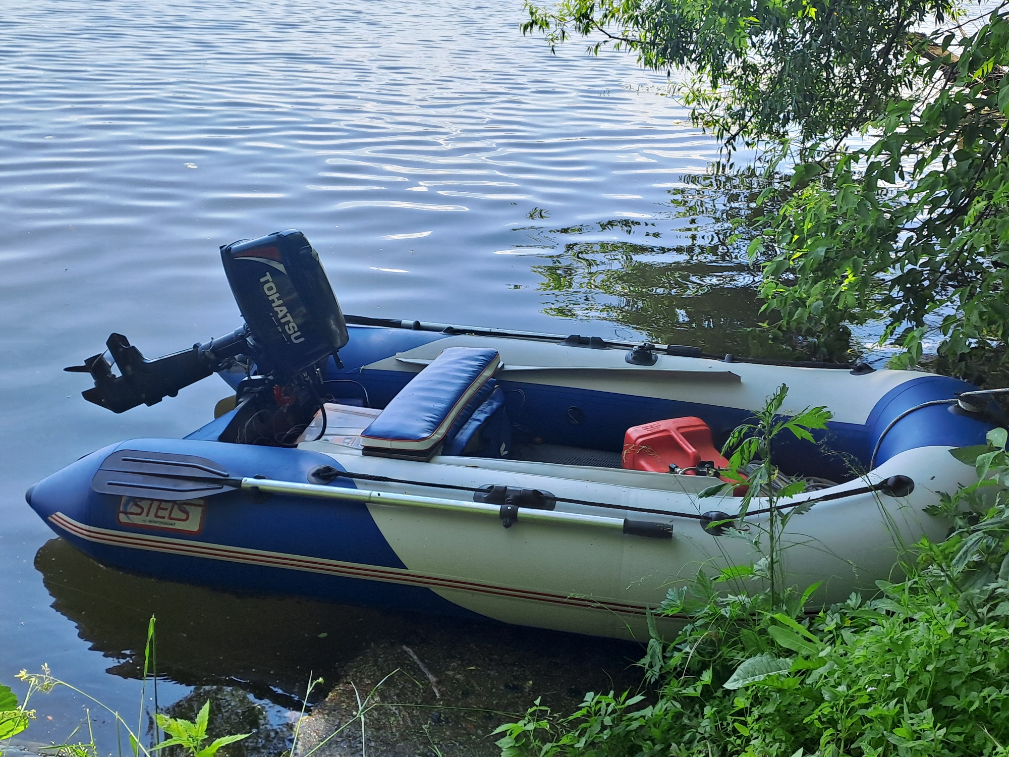 An inflatable boat with a Tohatsu outboard motor is docked on the shore of the Oka River in Dedinovo, Russia. The boat is blue and white with red stripes and has a cushioned seat. A red fuel can is visible inside the boat. Overhanging branches of a tree shade a portion of the water and boat, with green foliage growing along the bank. The water is calm with gentle ripples reflecting the sky and trees. The scene appears to be a tranquil, natural setting.