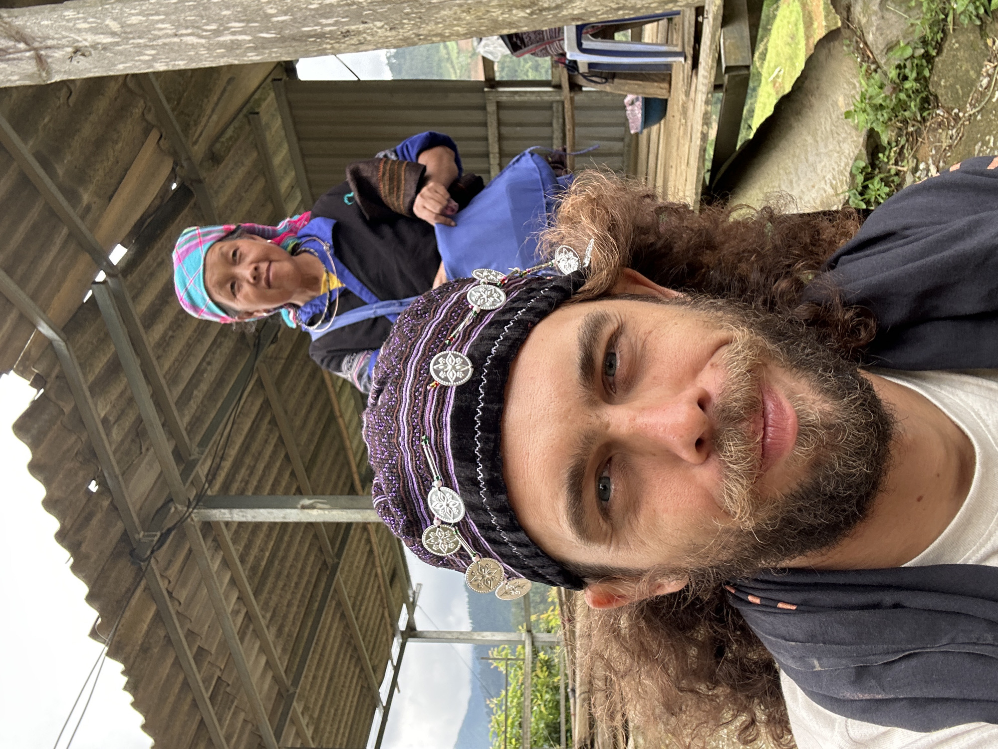 The image captures a close-up selfie taken in Dế Xu Phình, Vietnam, featuring a young adult male in the foreground and an elderly female in the mid-ground. The male, positioned in the lower left, has curly brown hair, a blonde-brown beard and mustache, and blue eyes, looking towards the camera with a slight smile. He wears a white t-shirt, a dark scarf, and a dark purple and black knitted cap embellished with multiple silver-colored medallions. In the upper right mid-ground, an elderly Asian female stands on a raised wooden platform, looking towards the camera. She wears a teal, pink, and purple plaid headscarf, a black jacket over a yellow shirt, silver hoop earrings, and a silver neck ring. She holds a blue fabric bag in her right hand. Overhead, a corrugated sheet metal roof is supported by metal beams and a weathered wooden post, with electrical wires running beneath. Distant green mountains and foliage are visible under an overcast sky in the background. On the wooden platform, a white and blue plastic chair and a blue bowl are present. The ground beneath the platform consists of rocks and small green plants.