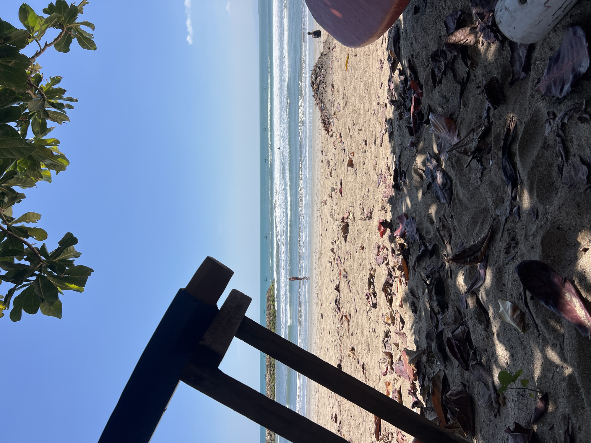 A low-angle, outdoor shot captures a tranquil beach scene in Kuta, Indonesia, under a clear blue sky. In the foreground, dry leaves are scattered across the sand, partially obscured by the shadow of a dark, wooden structure, possibly a beach chair or table. The middle ground reveals the sandy beach with gentle waves rolling onto the shore. A person is visible walking near the water's edge. Further out, several small boats dot the ocean, and a rocky breakwater extends into the sea. The background consists of the vast expanse of the ocean meeting the horizon. The overall atmosphere is serene and invites relaxation.