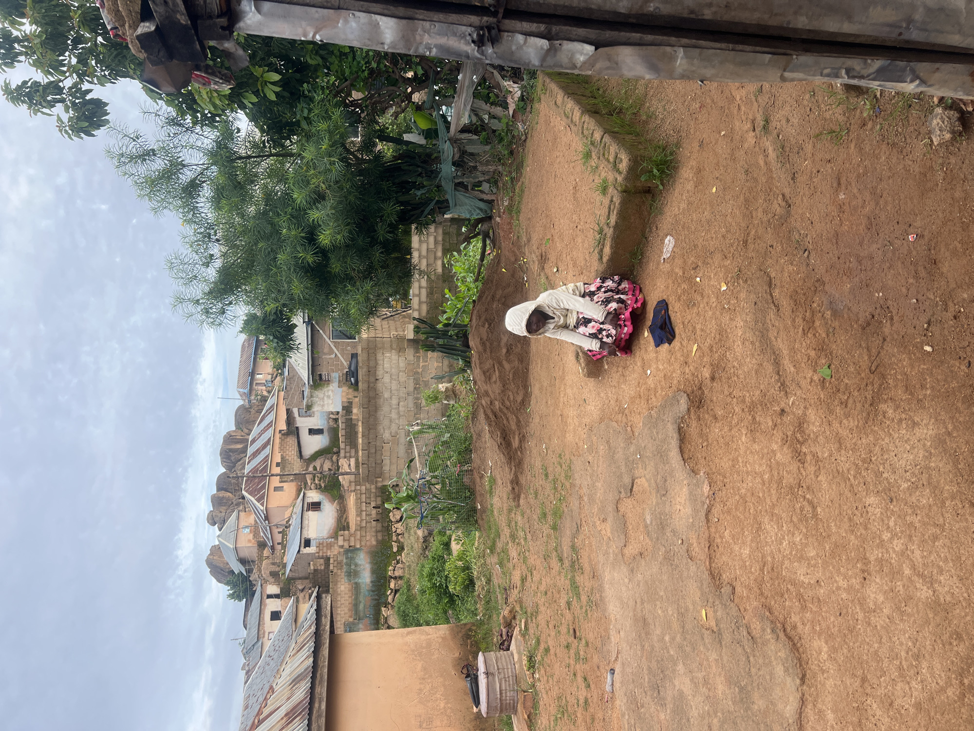 The image depicts a child in a white hoodie and a floral skirt, crouching on reddish-brown dirt in an outdoor setting, possibly a village or the outskirts of a town. The background features a cluster of houses with corrugated metal roofs and some brick structures, situated on uneven terrain with large rock formations visible further back. Dense green foliage, including trees and bushes, are interspersed among the buildings. In the foreground, there's a pile of dirt, a large metal container with a lid, and scattered debris. The sky is overcast. This scene is characteristic of the landscapes found in Jos, Nigeria, which is known for its rocky topography and mix of traditional and modern housing.