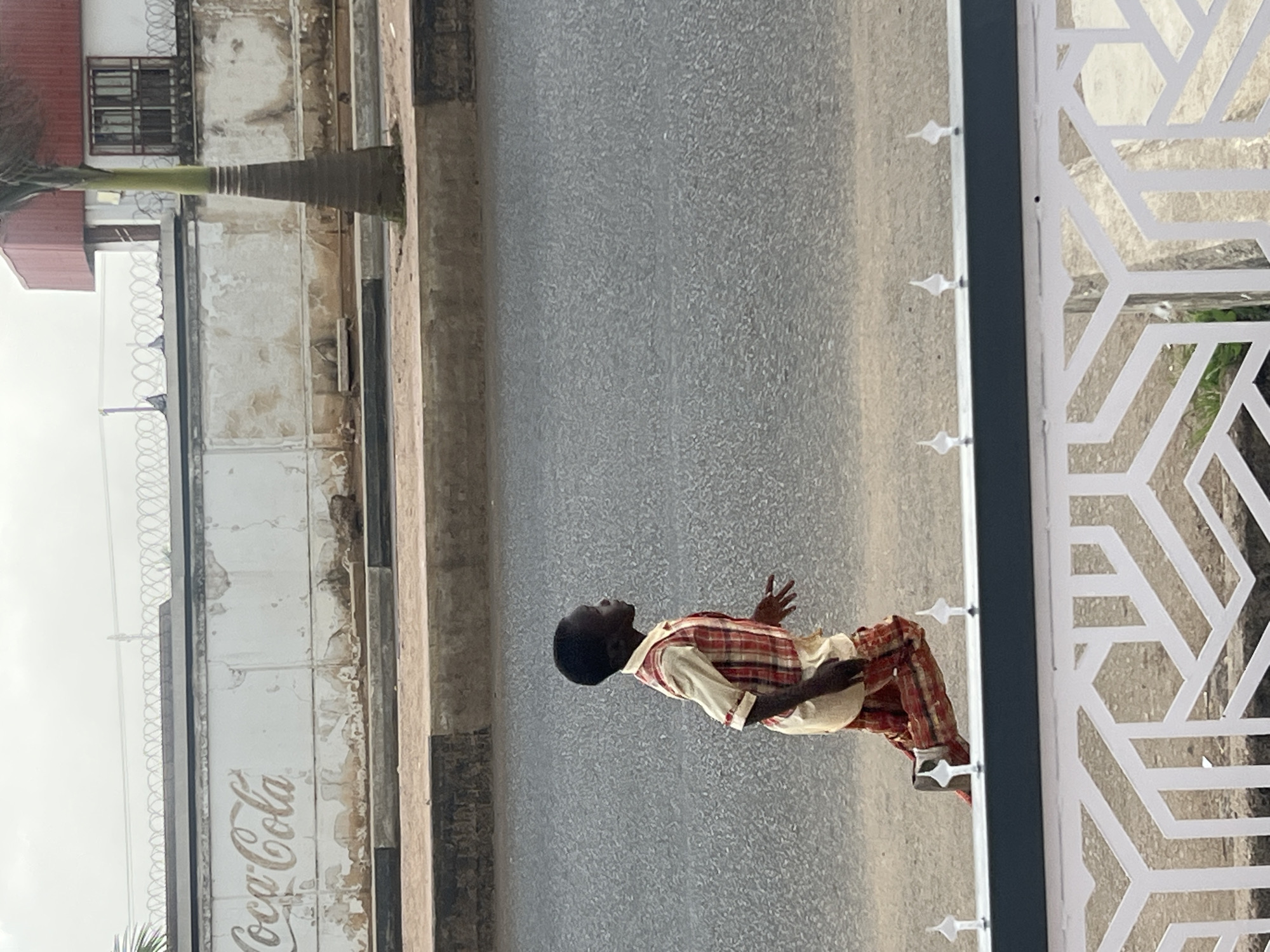 A young person is standing on a paved street in Barakin Lerawi, Nigeria. They are wearing a plaid shirt and tan pants, and are facing away from the camera, looking towards a building with a faded Coca-Cola advertisement on its wall. The building also has barbed wire along the top. A palm tree is visible to the left of the building. To the right of the person is a white, decorative fence.