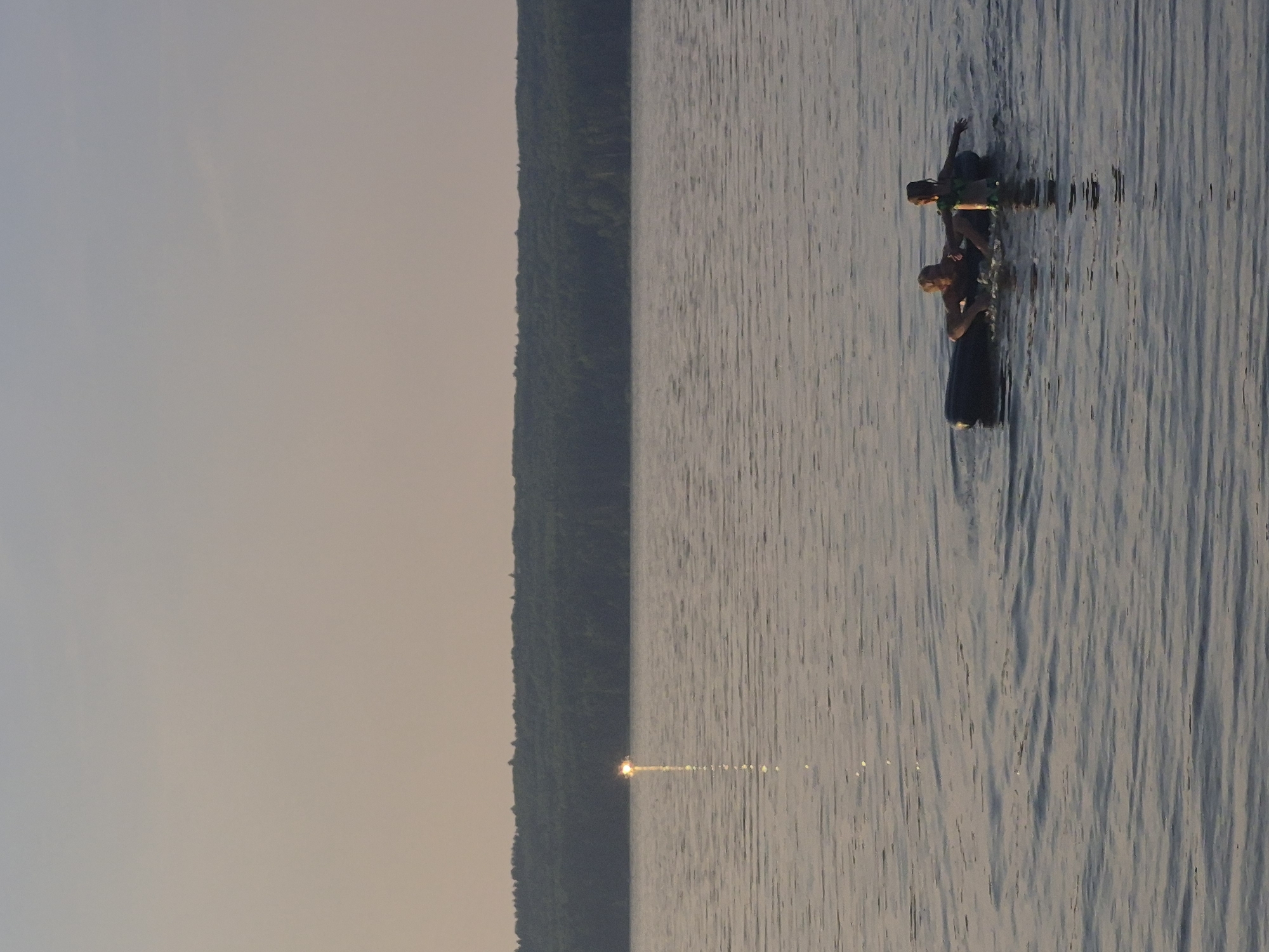 The media file depicts a serene scene on a body of water, likely a lake or river, near Chaikovsky, Russia. The time of day appears to be late afternoon or early evening, indicated by the soft, warm light reflecting on the water's surface and the hazy sky. The water is calm, with gentle ripples distorting the reflections.
The primary subjects are two individuals engaged in an activity on the water. They are positioned on what appears to be an inflatable raft or a similar floating device. One person is seated, while the other is standing and appears to be reaching out or adjusting their balance. Both individuals are wearing life vests, suggesting a focus on safety during their aquatic activity. Their interaction suggests a cooperative or playful moment, possibly fishing, enjoying recreation, or simply navigating the water.
In the background, a dense, dark line of trees forms a natural shoreline. This vegetation hints at a natural, possibly forested, environment. The contrast between the muted, hazy sky and the textured, reflective water creates a peaceful and somewhat melancholic atmosphere. The sun is low on the horizon, casting a shimmering golden path across the water, further emphasizing the time of day and the tranquil ambiance. There is no visible text in the media file.