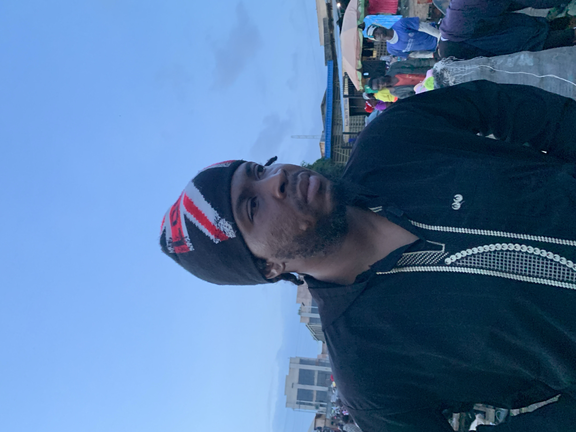 A medium shot captures a man wearing a black beanie with a Union Jack pattern and a black collared shirt with white embroidered detailing. He has a beard and is looking to his right, with his mouth slightly open. The background depicts a bustling outdoor market scene. Several stalls are visible, with people browsing and interacting. Buildings, possibly shops or residences, line the background, suggesting an urban environment. The sky is overcast with a pale blue hue. The image appears to be taken in Gura Top, Nigeria, indicated by the context of similar images from the area.