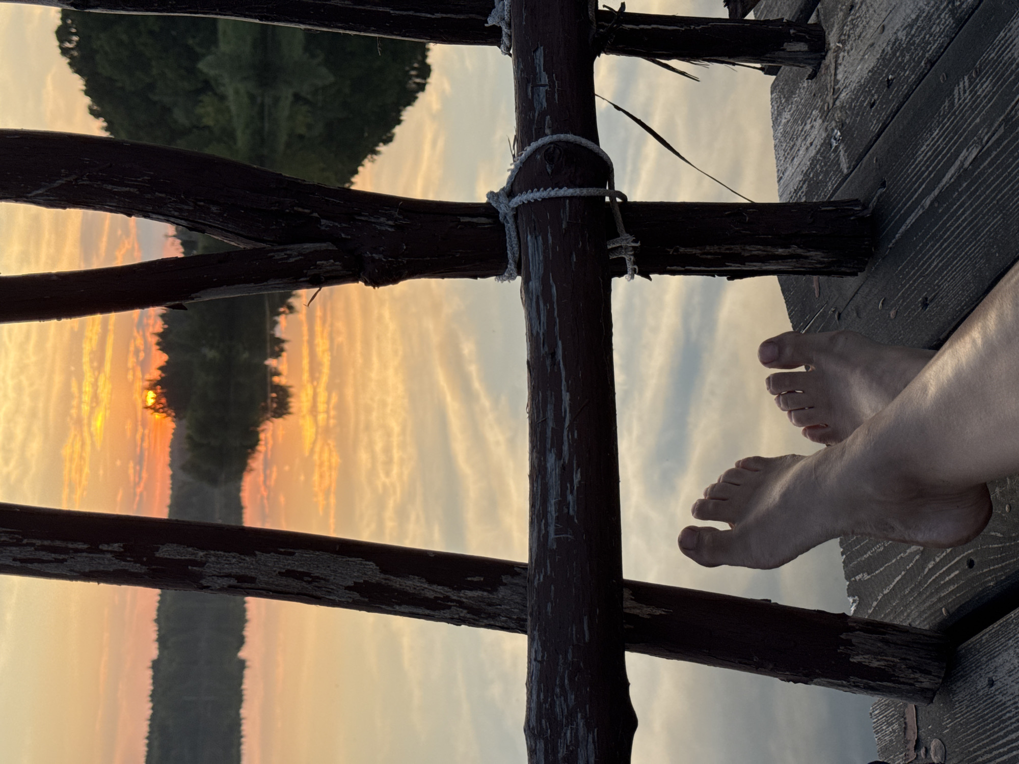 The image shows a close-up view of a person's bare feet resting on a wooden platform, with a serene sunset reflected in a body of water in the background. The wooden platform appears to be part of a dock or pier, with rustic wooden posts and a weathered railing visible. The railing is secured with rope in some areas. The sunset casts a warm glow across the sky and water, with shades of orange, yellow, and pink. The reflection shows clouds and the silhouetted outline of trees and a distant shoreline. The image is taken from a low angle, focusing on the feet and the foreground of the platform. The specific location in the city of Kamysh, Russia, is not discernible from the visual information presented in the image.