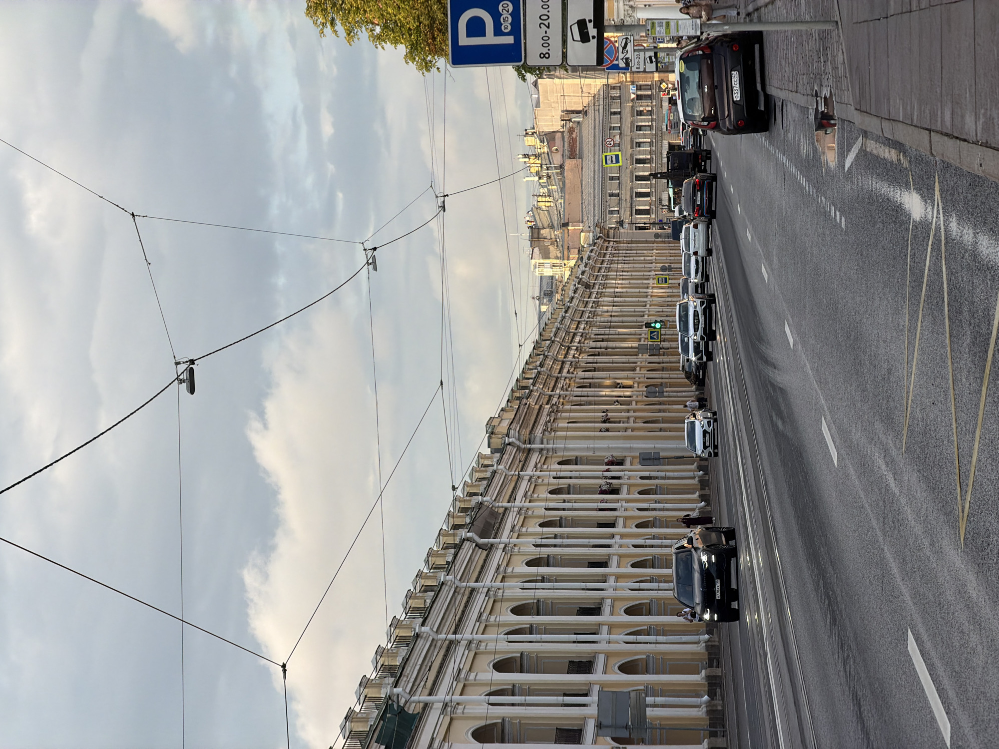 The image depicts a wide, asphalt street in Saint Petersburg, Russia, likely on a cloudy day. The street is lined with classical architecture characterized by a long building facade with arcades and columns on the left. Numerous cars are present on the road, including a black SUV in the foreground, a white sedan, and other vehicles further down the street. Overhead, a complex network of wires for what appears to be a tram or trolleybus system stretches across the sky. On the right side of the street, there is a sidewalk with a parking sign indicating restrictions from 8:00 to 20:00. A few pedestrians are visible on the sidewalk, and a brown Kia Soul is parked. The overall scene suggests an urban environment with active traffic and historical architectural elements typical of Saint Petersburg.
