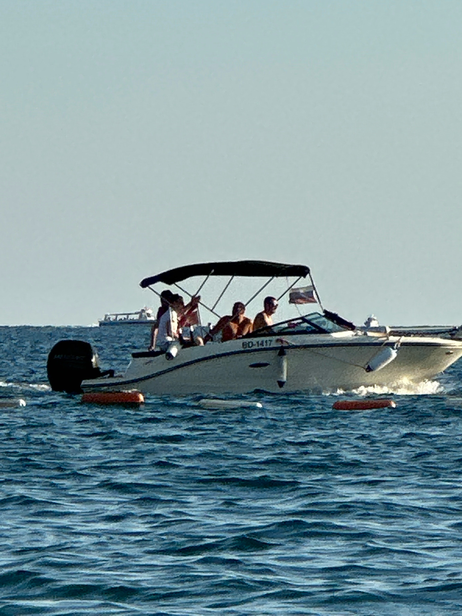 The image depicts a sunny day at sea, likely off the coast of Boreti, Montenegro. In the foreground, the choppy blue water is disturbed by the wake of a white speedboat. The speedboat is moving from left to right, creating white foam. Several people are visible on the boat, including a person steering, and at least three passengers. The boat has a black canopy providing shade. A flag is flying from a pole at the back of the boat. In the mid-ground, a larger ship or ferry with multiple decks is visible on the horizon. The sky is a clear, pale blue. The overall scene suggests recreational activity on the water.