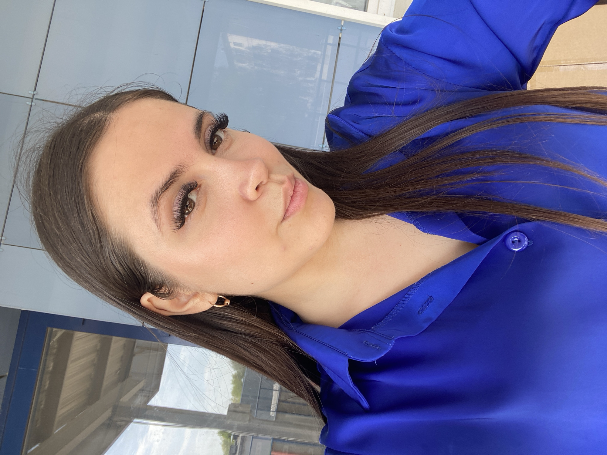 A woman with long brown hair, fair skin, and full eyelashes is wearing a bright blue button-down shirt. She is positioned in front of a modern building with reflective windows and gray paneling. An earring is visible on her left ear. The location may be Chaikovsky, Russia, but this is not visually confirmed. The focus is on the woman's face and upper body. The overall setting appears urban and contemporary.