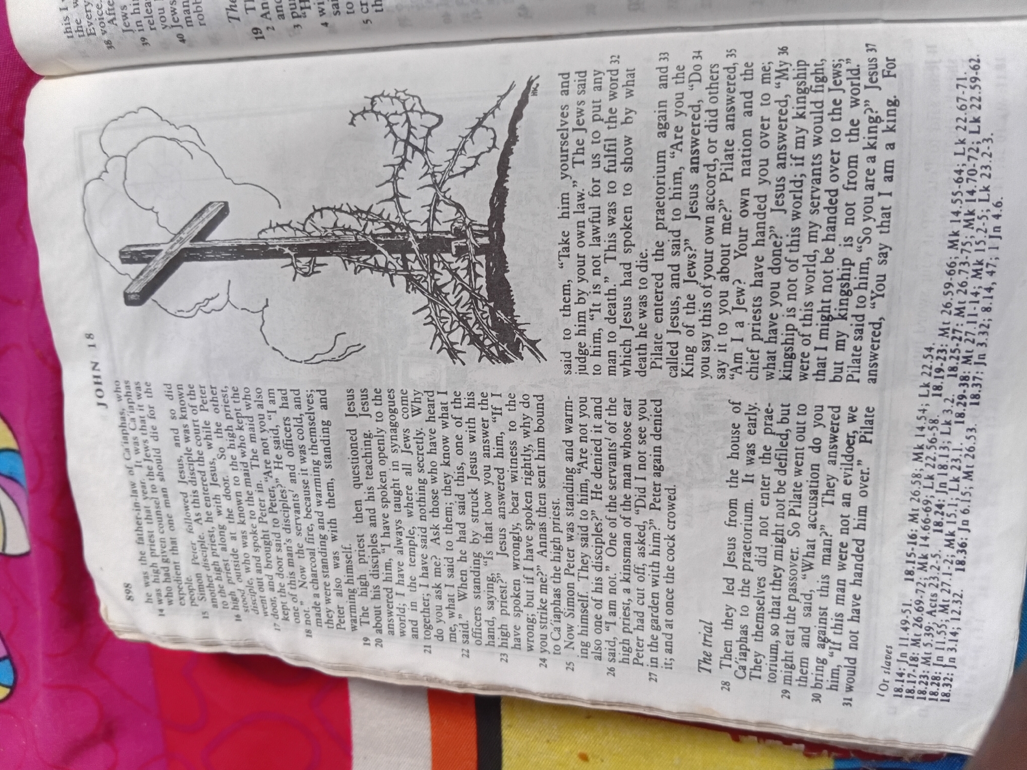 The image shows an open book, specifically a Bible, displaying a page from the Gospel of John, chapter 18. The page features an illustration of a cross adorned with a crown of thorns, set against a backdrop of stylized clouds. The text on the page is in English and includes a verse-by-verse breakdown of the biblical narrative, detailing events related to Jesus' trial.
The text describes Jesus' questioning by the high priest, Peter's denial of Jesus, and the subsequent events leading to Jesus' appearance before Pilate. Key figures mentioned include Jesus, Peter, Annas, Caiaphas, and Pilate. The passage covers the time of day, with "It was early" noted for Jesus' leading to the praetorium.
The Bible itself is open to page 898, with "JOHN 18" clearly visible. There are also cross-references to other books of the Bible and their corresponding verses printed in smaller font at the bottom of the page. The overall presentation suggests a religious context, likely a personal study or devotional moment. The book appears to be lying on a pink textured surface.