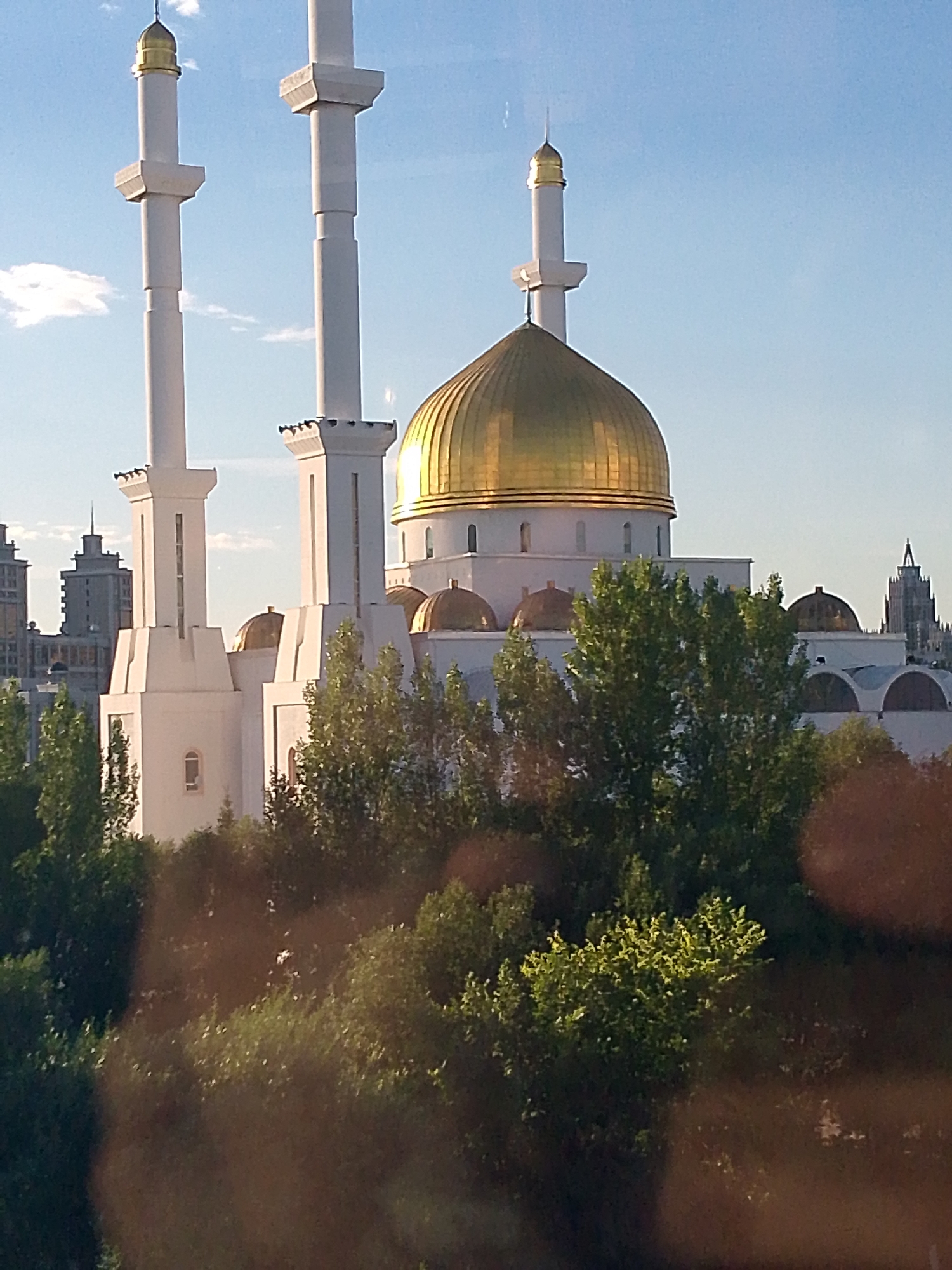 The image shows the Hazrat Sultan Mosque in Astana, Kazakhstan. The mosque features a large golden dome and two tall white minarets, which are characteristic of Islamic architecture. The mosque is surrounded by lush green trees, and in the background, modern city buildings and a clear blue sky can be seen. The lighting suggests it is daytime, possibly late afternoon, given the angle of the sun. The photo appears to be taken from a distance, possibly from another building. There are no people visible in the image.