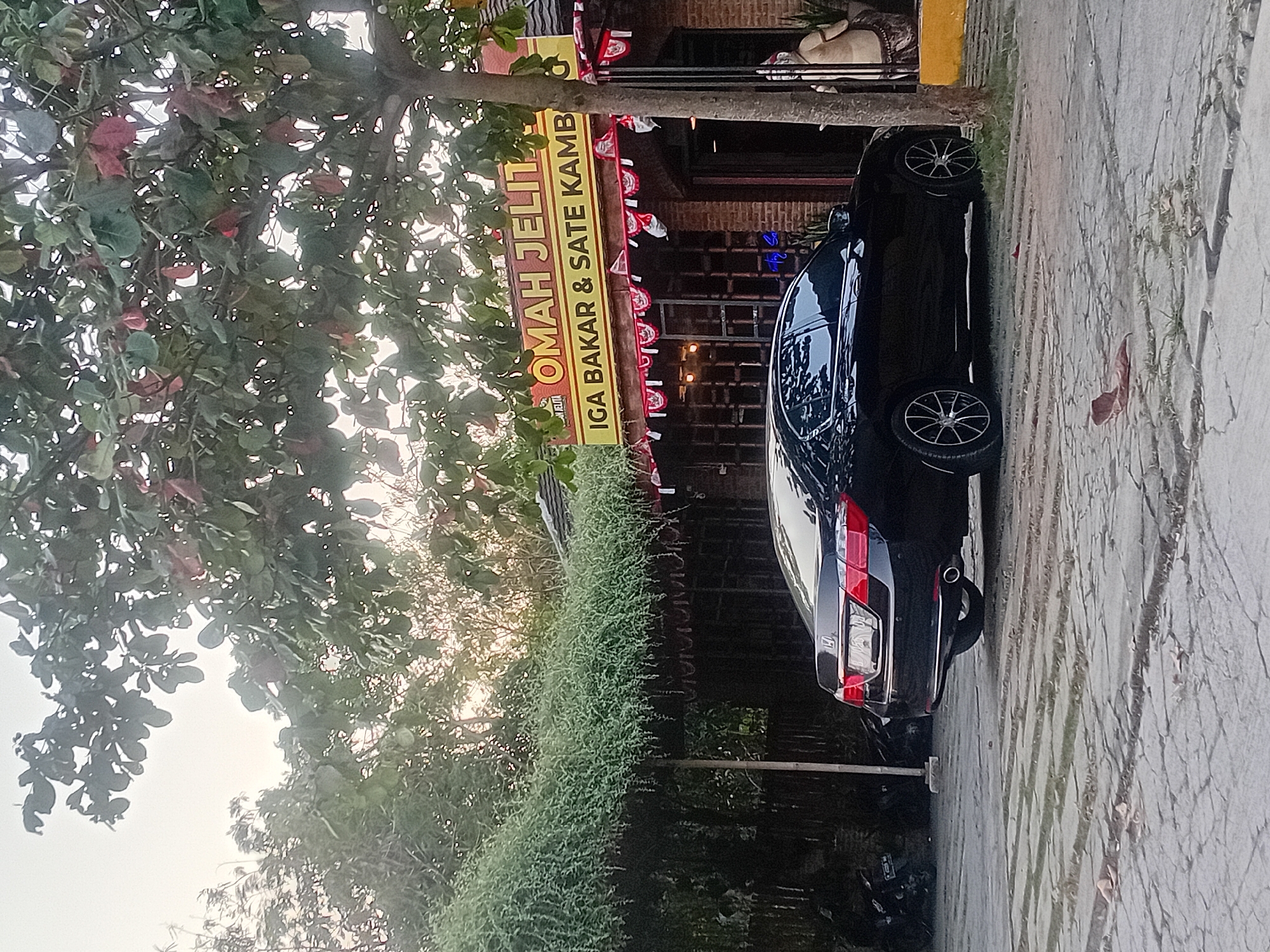 A black sedan is parked on a paved outdoor area in front of a building with a yellow sign that reads "OMAH JELIT" and "IGA BAKAR & SATE KAMB". The building has red brick walls and is decorated with red and white pennant banners. Lush green foliage hangs over the building's entrance, and a tree with green and reddish leaves stands to the right of the frame. The ground is a textured gray pavement. In the background, there is a hint of a motorcycle and a statue. The sky is overcast and bright. The scene appears to be in Bae, Indonesia.