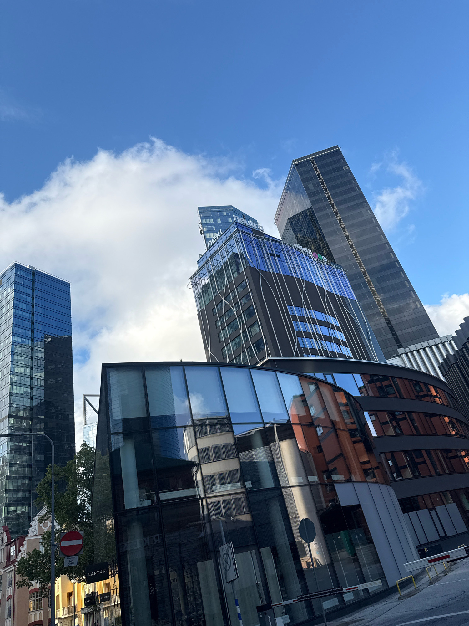 The image captures a modern architectural landscape in Tallinn, Estonia, focusing on the sky-high buildings and their reflections. The scene is dominated by a clear blue sky with scattered white clouds.
In the foreground, a contemporary building with a curved, glass facade reflects the surrounding structures and the sky. The glass displays distorted images of buildings, including one with reddish-orange brickwork and windows. Several barriers and a street lamp are also visible in this section.
Further back, a cluster of tall, modern office buildings forms the city's skyline. To the left, a blue glass skyscraper stands prominently. Adjacent to it, a darker, taller skyscraper with a reflective dark facade rises into the sky. In the center, another building features a dark exterior with striking blue glass sections on its upper floors, adorned with white vertical lines and the "nexi" logo.
The lower parts of the image show glimpses of older architecture with traditional windows and decorative elements, juxtaposed against the sleek, modern designs. A circular red and white traffic sign with a bicycle symbol is mounted on a street lamp. The street level is partially visible, with some pavement and barriers. The overall impression is one of urban development and modern architecture in a city setting.