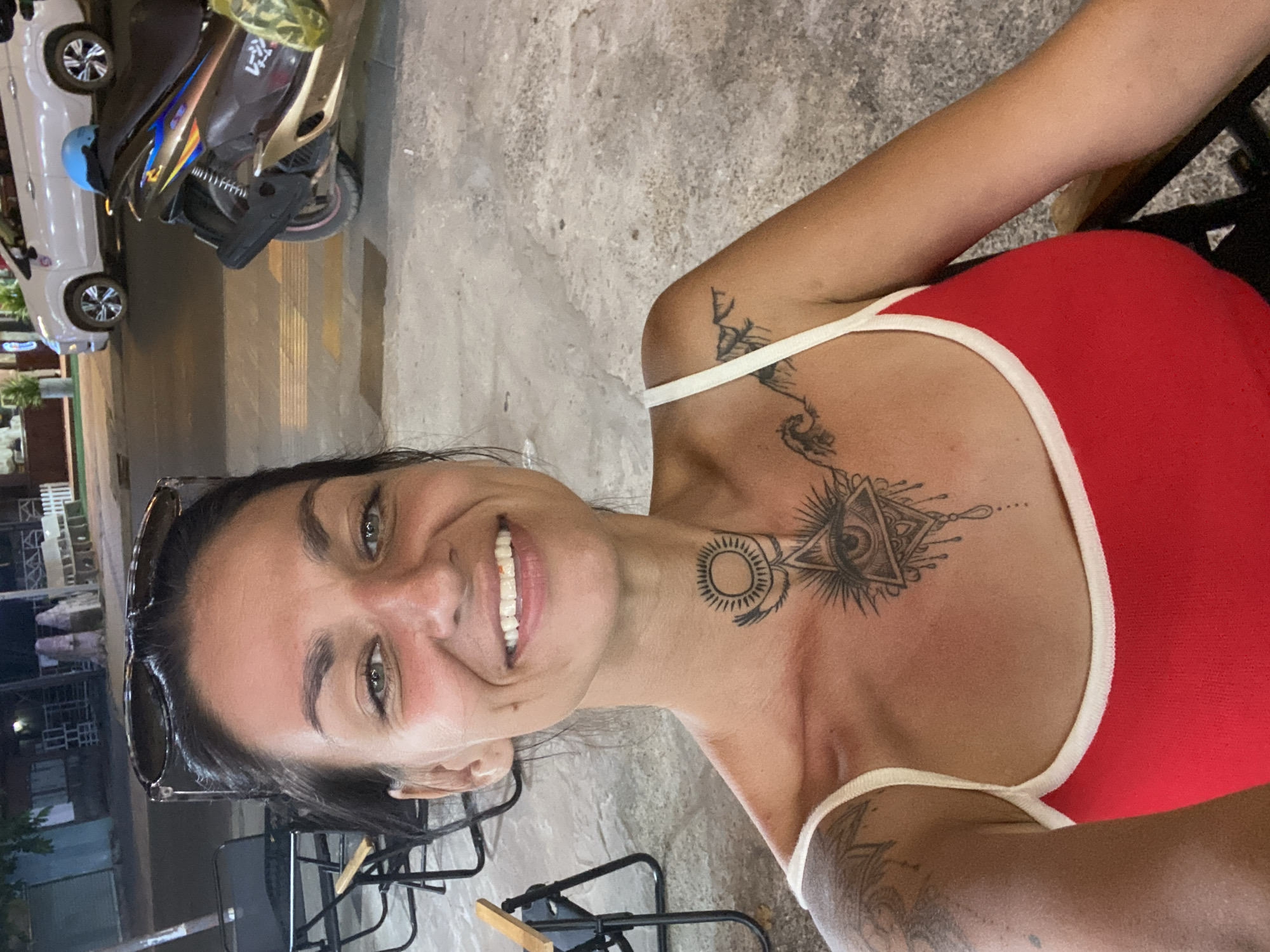 A woman with dark hair, wearing sunglasses pushed up onto her head and a red top with white trim, smiles at the camera. She has several tattoos: one on her upper chest depicting an eye within a geometric design, another on her neck of a sun-like symbol, and more tattoos visible on her arm. She is seated, and the image is taken from a high angle.
In the background, a white car and a brown and gold scooter are parked. The scooter has a yellow and green bag on the back. The scene appears to be outdoors, possibly on a street or sidewalk, with concrete ground. The lighting suggests it is evening or nighttime.
The location context is Phu Thuy Ward, Vietnam.