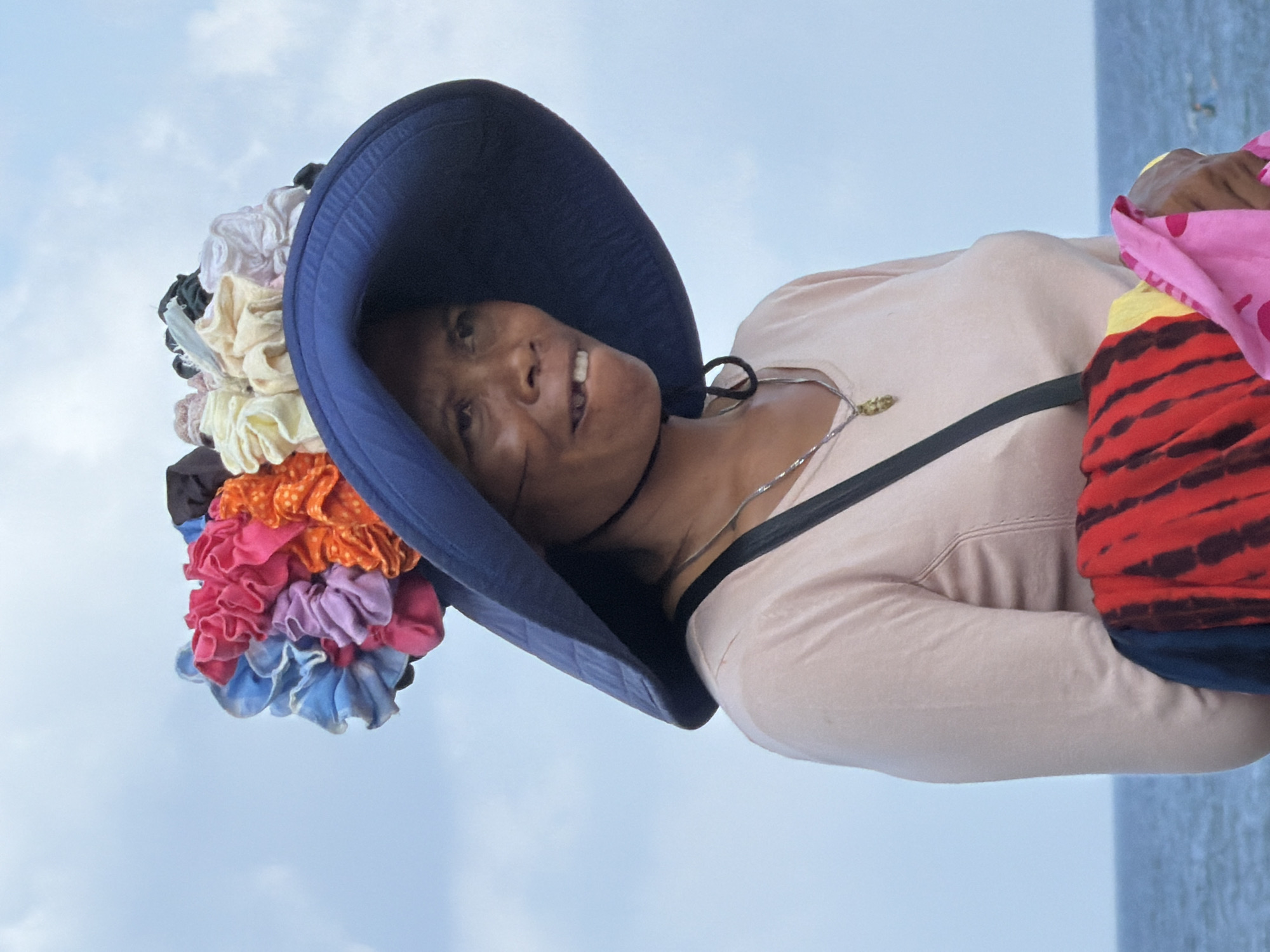A medium-shot portrait shows an East Asian woman in a broad-brimmed blue hat adorned with an assortment of colorful fabric flowers. She wears a light pink long-sleeved shirt and a silver necklace with a pendant. Her gaze is directed to the right, and her mouth is slightly open, revealing a missing front tooth. The background consists of a pale blue sky with scattered clouds and the ocean in the distance. The woman is holding a bundle of vibrant, tie-dyed fabric, predominantly red and yellow, along with a pink fabric with polka dots, suggesting she may be a vendor or engaged in trade near a body of water, possibly on the coast of North Kuta, Indonesia.