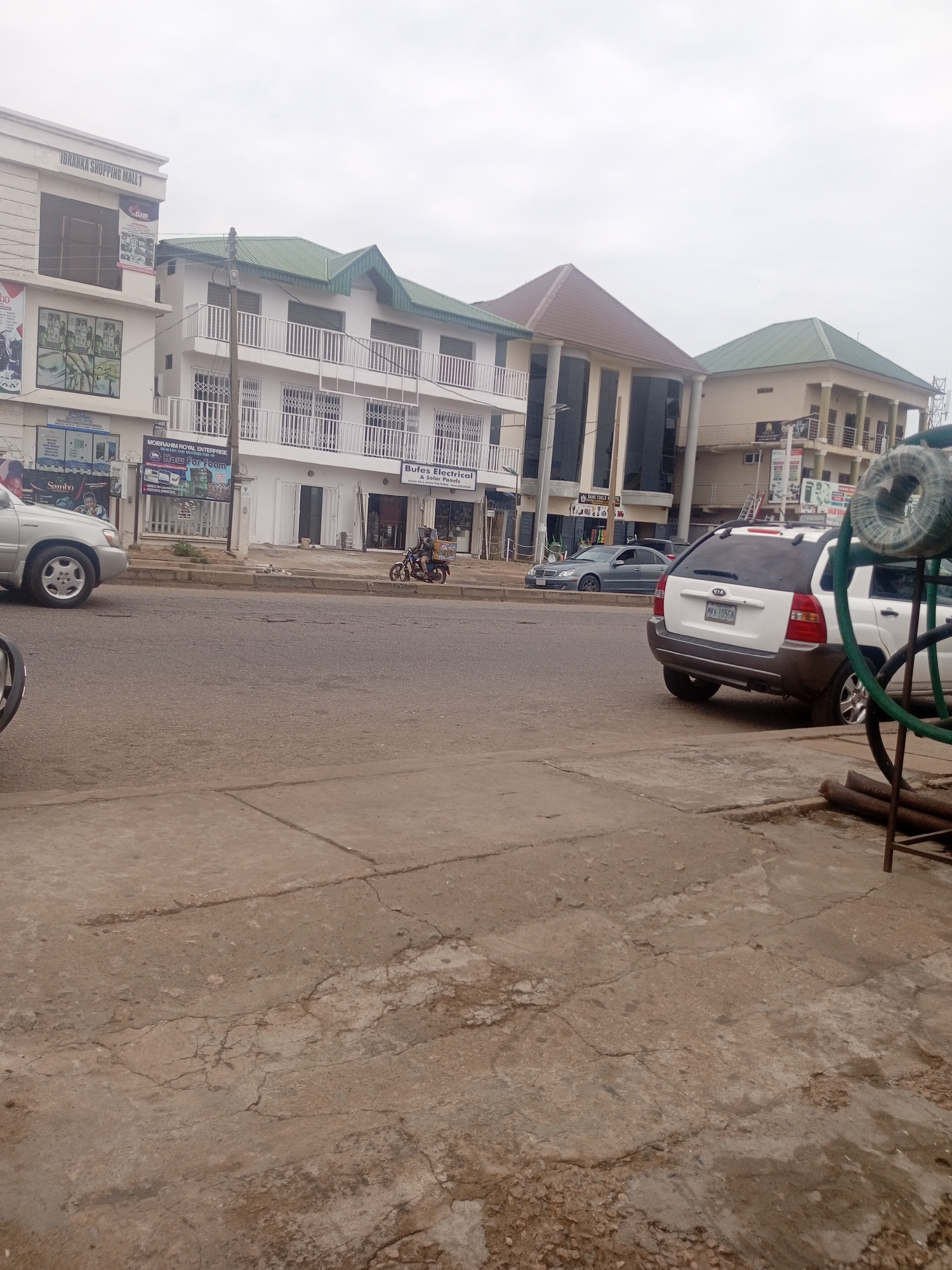In Sardauna, Nigeria, an overcast sky looms over a street scene. On the right, a white Kia Sportage SUV with license plate MKK-105GN is parked on the road. To its left, a silver sedan is moving along the asphalt road. Further left, a silver car is parked at the curb in front of a building. A person on a motorcycle is also visible on the road. The buildings lining the street feature modern architecture with white exteriors and green roofs. Signs indicate businesses such as "IBRAHIM SHOPPING MALL 1," "Bufes Electrical & Solar Panel," and "MOIBRAHIM ROYAL ENTERPRISE." A stack of coiled green material, possibly hoses or wiring, rests on the ground in the foreground on the right. The road is made of asphalt with visible cracks and patches, and a concrete sidewalk is present on the right side of the image.
