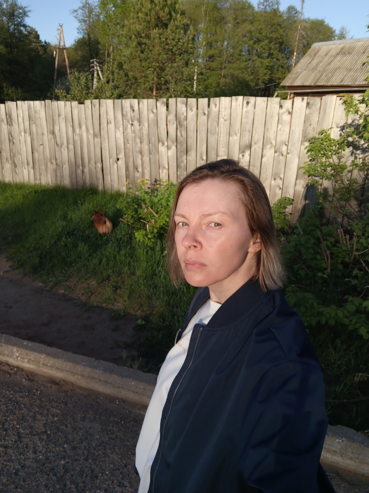 A woman with short brown hair, wearing a white t-shirt and navy bomber jacket, is standing outside. Behind her is a wooden fence, green grass, vegetation, a dog, and a house. Utility poles are visible in the background. The photograph was taken in Chaikovsky, Russia.