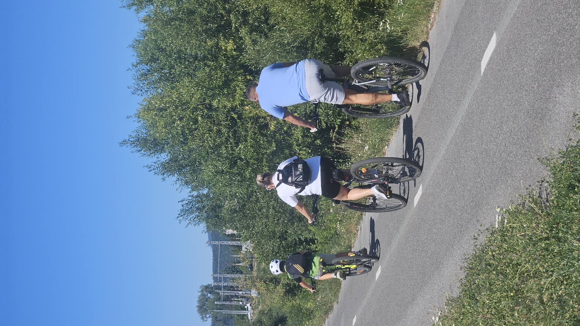 Three people, a child and two adults, are cycling on a paved path in Prešov, Slovakia. The child is in the foreground, wearing a white helmet and a green and black shirt, riding a bicycle with yellow accents. Behind the child is an adult woman in a white t-shirt and black shorts, with a black backpack. Further back is an adult man in a light blue t-shirt and gray shorts, also on a bicycle. The path is bordered by green vegetation and trees on the right side, and a grassy verge with some wildflowers on the left. In the background, there are power lines and a distant hillside. The sky is clear and blue.