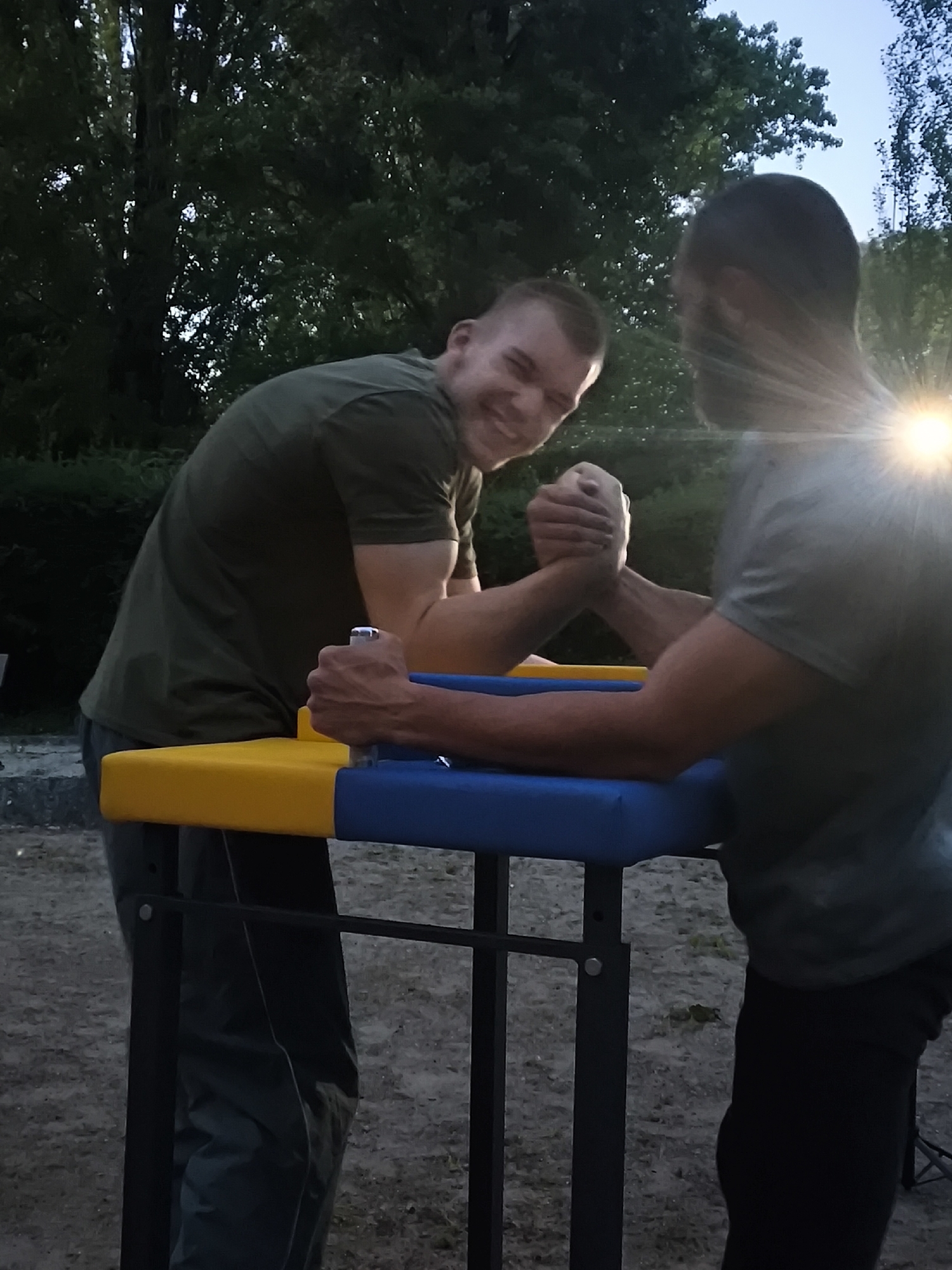 Two men are engaged in an arm wrestling match on a specialized table outdoors. The man on the left, wearing a dark green short-sleeved shirt and dark trousers, is seen from the chest up, smiling broadly and looking towards the camera. His left arm is locked in a grip with the other man's right arm, and his right hand is holding a yellow peg on the arm wrestling table. He appears to be leaning into the match with effort or enthusiasm.
The second man, on the right, is viewed from the back and side, wearing a grey short-sleeved shirt and dark trousers. His face is not visible, but his right arm is engaged in the arm wrestling grip, and his left hand is holding a silver-colored peg on the blue padded side of the table.
The arm wrestling table features distinct yellow and blue pads and a black metal frame. The ground beneath them is a dirt or gravel surface. In the background, there are numerous dark green trees, suggesting an outdoor, natural setting. The sky is partially visible, appearing bright blue in some areas, hinting at a clear day. A strong sun flare is prominent behind the head of the man on the right, indicating that the sun is low in the sky, possibly late afternoon or early evening. The overall atmosphere appears to be one of friendly competition. The location context is Schiltigheim, France. No visible text is present in the image.