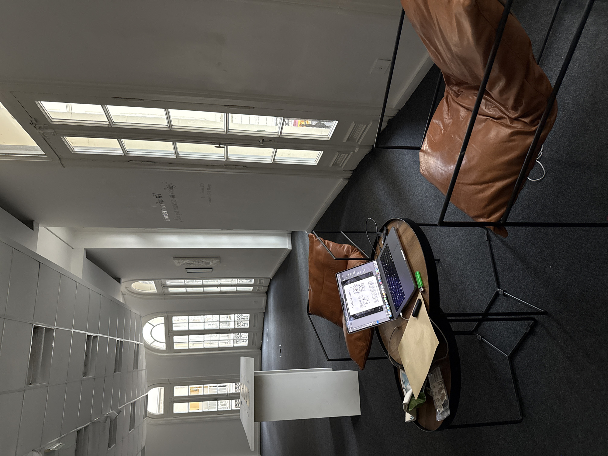The image depicts the interior of an office space, likely in Buenos Aires, Argentina. The space features several large windows with visible mullions. A white wall displays the text "Conversation #1" and "(Qué vas a hacer con el lago?)".
Two chairs are visible; one upholstered in brown leather is situated to the right, and another, also upholstered in brown leather, is placed at a small, round table. On the table is a laptop. A paper bag with items, possibly trash, is next to the laptop.
Further down the space, more windows can be seen. A white rectangular object, possibly a counter or a desk, is visible. The floor appears to be covered with dark grey or black carpet. The space is well-lit, likely from natural light entering through the numerous windows.