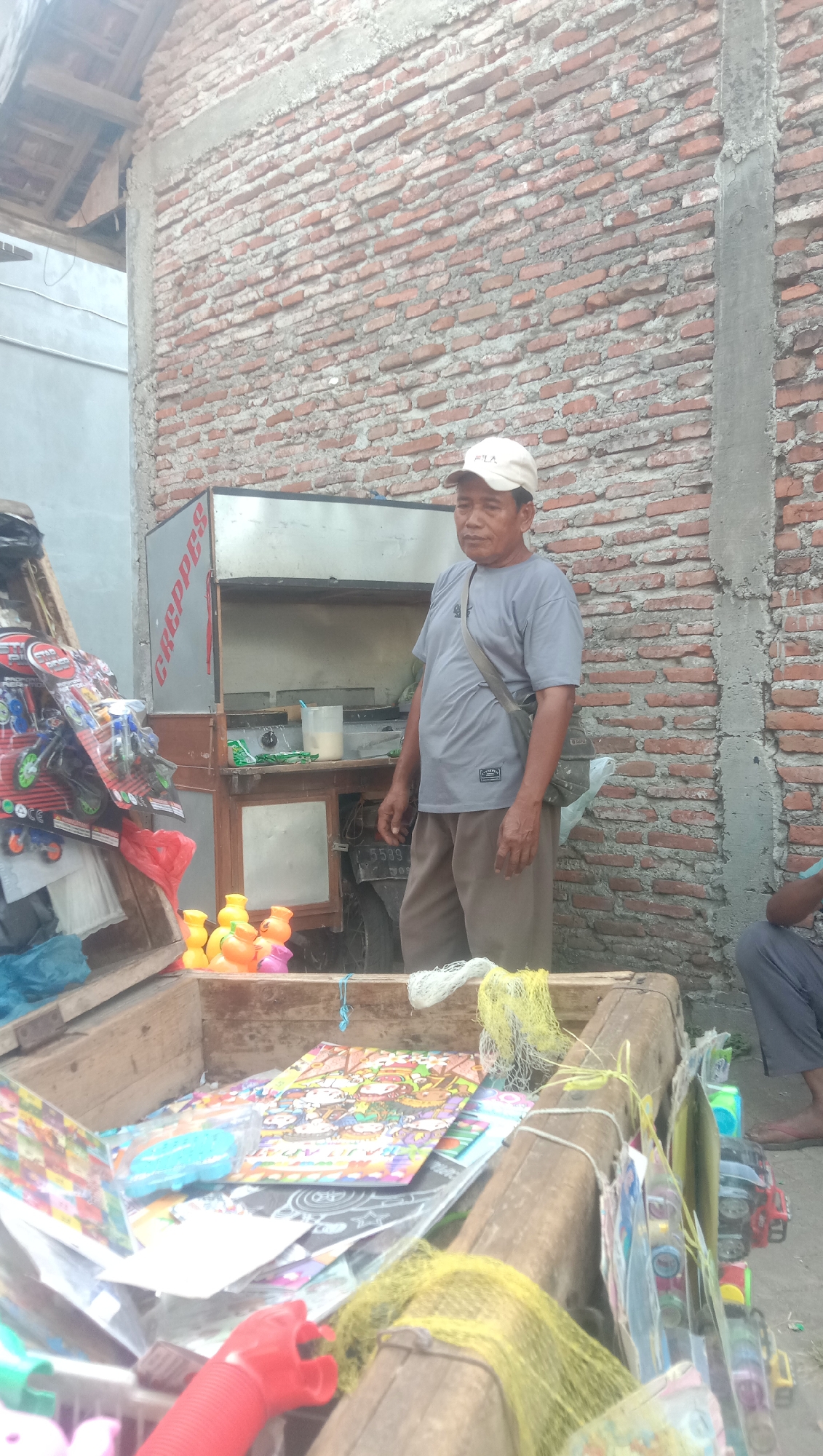 A man stands behind a crepes cart in Karanganyar, Indonesia. He is wearing a grey t-shirt, brown pants, and a white baseball cap. A grey bag is slung across his chest. The crepes cart is metallic and has the word "CREPPES" written on its side. In front of the cart, there is a wooden cart filled with various items, including colorful toys, what appear to be stickers or posters, and some yellow mesh netting. A motorcycle is parked behind the crepes cart. The background consists of a brick wall and a tiled roof. The overall scene suggests a street vendor setup in a casual, outdoor environment.