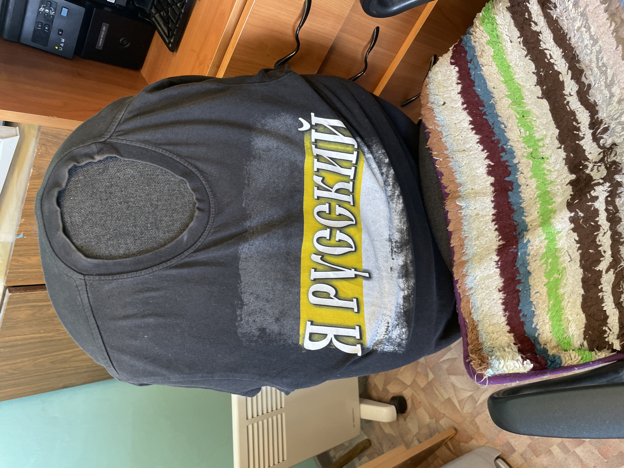 The image shows a black t-shirt with the Russian text "Я РУССКИЙ" (I am Russian) printed vertically on it. The shirt is partially draped over a striped seat cushion on a chair. The environment appears to be an indoor setting, possibly a home or office. Details visible include a printer on a wooden surface, and a radiator. There is no verifiable information in the image about the location in Sarapul, Russia.
