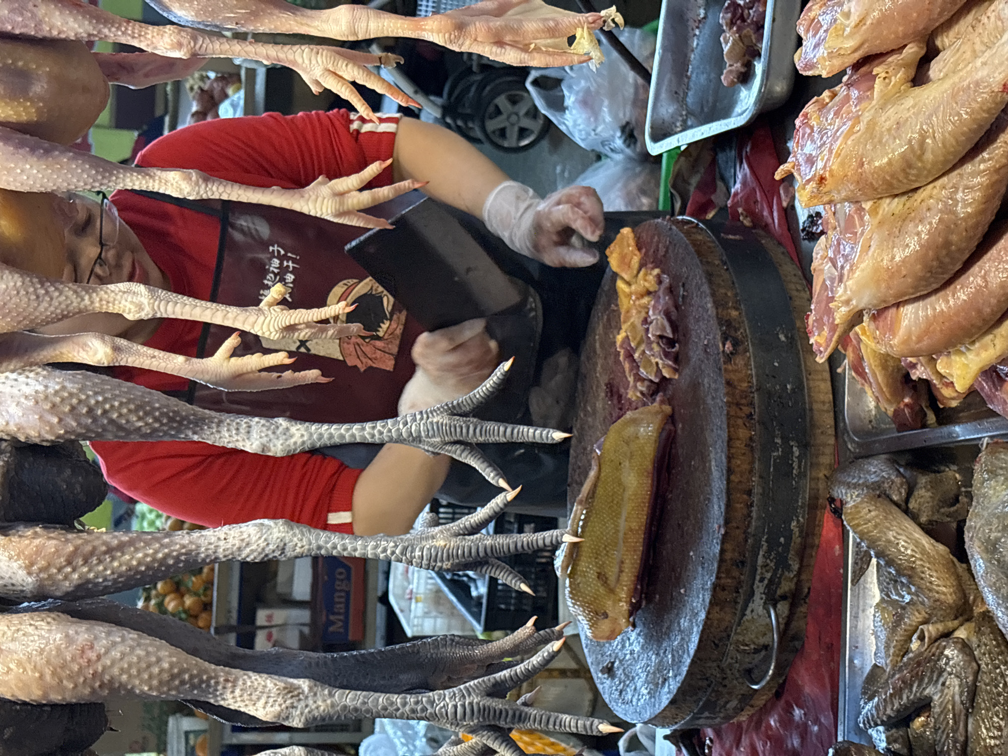 The image depicts a market scene, likely in Jiangjin, China, featuring a butcher preparing poultry. The butcher, wearing a red shirt, apron, and gloves, is using a cleaver on a large wooden cutting board. Various poultry parts, including chicken feet, whole birds, and cut pieces, are visible on and around the workspace. The environment suggests a bustling market, with a mix of colors, textures, and potential commercial activity.