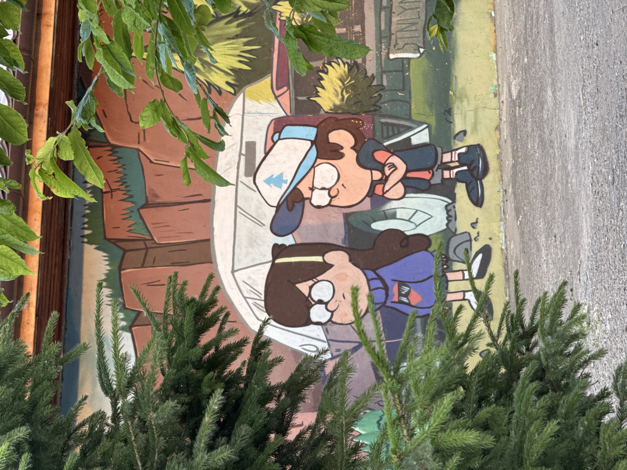 This media file features a mural painted on a wall, depicting cartoon characters in a somewhat stylized, perhaps slightly unsettling, artistic style. The mural is partially obscured by foliage, with green leaves from a tree hanging down from the top and evergreen branches in the foreground. The scene shows two young characters, appearing to be children, rendered in a bold, animated style. One character, to the upper left, wears a blue cap with a light blue brim and a symbol that resembles a stylized arrow or pine tree. This character has brown hair and is wearing a dark blue shirt with orange accents. The other character, to the lower right, has brown hair and wears glasses. They are dressed in a purple top with an orange and black emblem. The characters appear to be lying down or reclining, with their faces turned towards the viewer. The background of the mural includes elements that suggest an outdoor setting, with a textured brown area resembling rock or earth, and patches of green and yellow, perhaps representing foliage or a landscape. There is a hint of a sign in the background with some text, but it is difficult to decipher clearly due to its stylized nature and the angle. The overall impression of the artwork is somewhat playful, but the slightly distorted features of the characters might evoke a sense of unease for some viewers. The presence of natural elements like leaves and branches suggests the mural is located outdoors, possibly in a park or an urban area with some greenery. The lighting suggests it is daytime, with the sun casting some shadows.