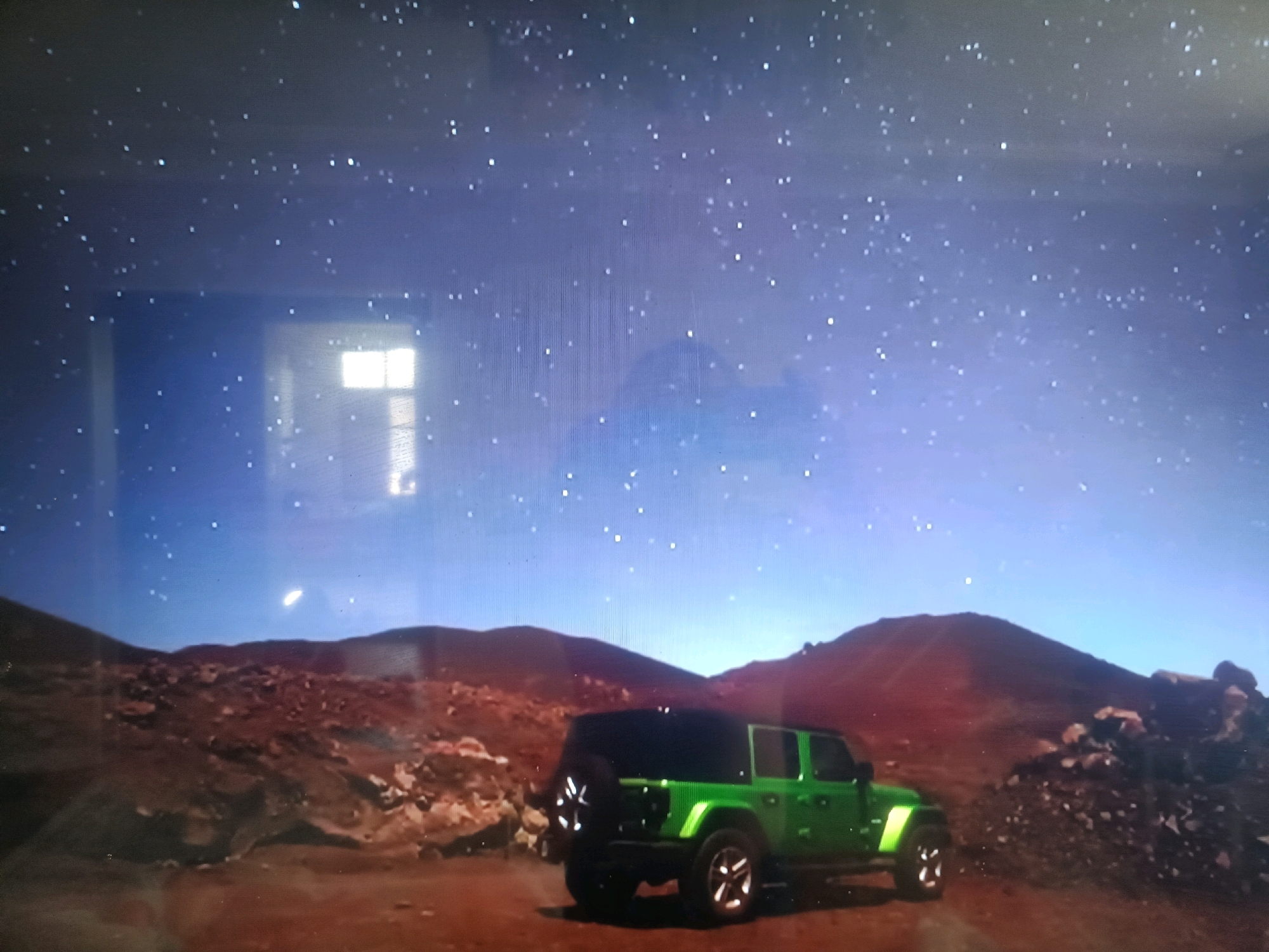 The image depicts a striking scene under a vast, starry sky. In the foreground, a vibrant green Jeep Wrangler is parked on a rocky, reddish-brown terrain. The vehicle, a four-door model with tinted windows and a spare tire mounted on the rear, is positioned on a dirt path or road. The surrounding landscape is arid and mountainous, with rugged, dark-colored hills and scattered rocks. The sky is a deep blue, filled with numerous bright stars, and a faint glow suggests the presence of artificial light or perhaps distant celestial bodies. There are no people visible in the image. The overall impression is one of isolation and adventure in a rugged, natural environment. The location is specified as Қасымбек датқа, Kazakhstan, which is likely a rural or mountainous area given the landscape shown.