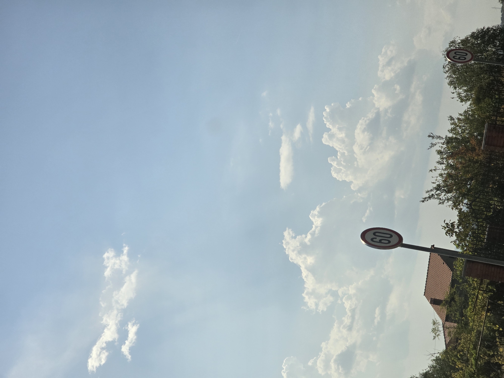 The image depicts a clear blue sky with scattered white cumulus clouds. In the foreground, on the left, a red-tiled roof of a house is visible above a dense canopy of green trees. To the right of the house and slightly forward, a speed limit sign indicating "60" is prominently displayed on a metal pole. Further to the right and slightly behind the first speed limit sign, another identical "60" speed limit sign is partially visible. The signs are circular with a white background, a red border, and black numerals. The environment appears to be an outdoor setting, likely in a suburban or semi-rural area, given the presence of a house, trees, and traffic signs. The lighting suggests it is daytime, with the sun illuminating the clouds and casting subtle shadows. The location is Podgorica, Montenegro.