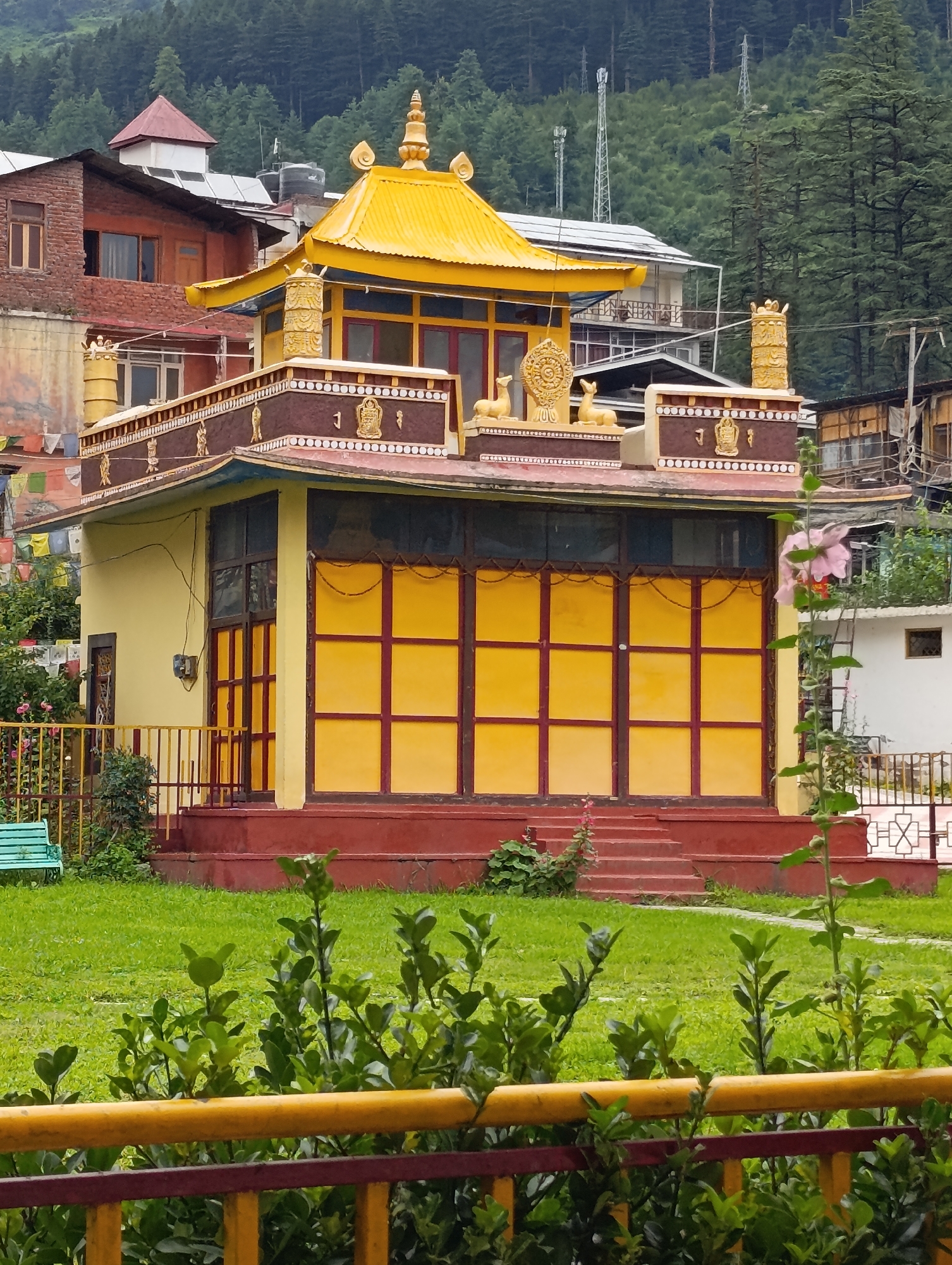 The image shows a traditional Buddhist monastery in Manali, India. The monastery is a two-story building with a yellow and red color scheme. The lower story is painted yellow with large red-framed windows. The upper story has a decorative frieze with golden figures and symbols, and a bright yellow roof topped with a golden spire. The monastery is situated on a grassy slope, with trees and buildings visible in the background. A pink flower is in bloom on the right side of the image. The scene suggests a peaceful and serene atmosphere.