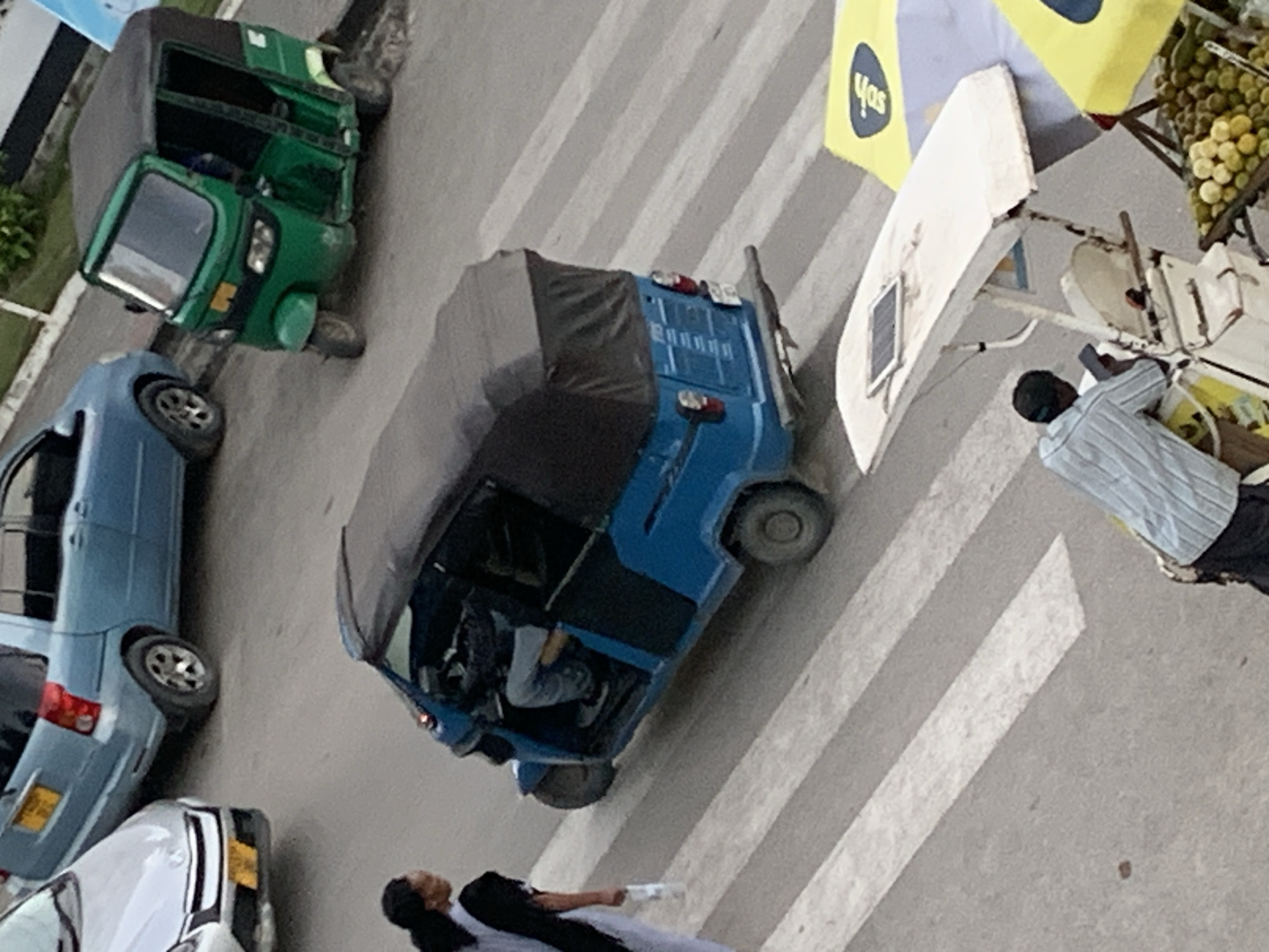 The image shows a street scene in Dar es-Salaam, Tanzania from an elevated perspective. A green auto rickshaw, a blue auto rickshaw, and two cars are parked on the left side of the street. A man in a striped shirt stands near a fruit stand with a yellow umbrella. A woman in a black top and white dress walks across a crosswalk. The street is paved and marked with crosswalk stripes.