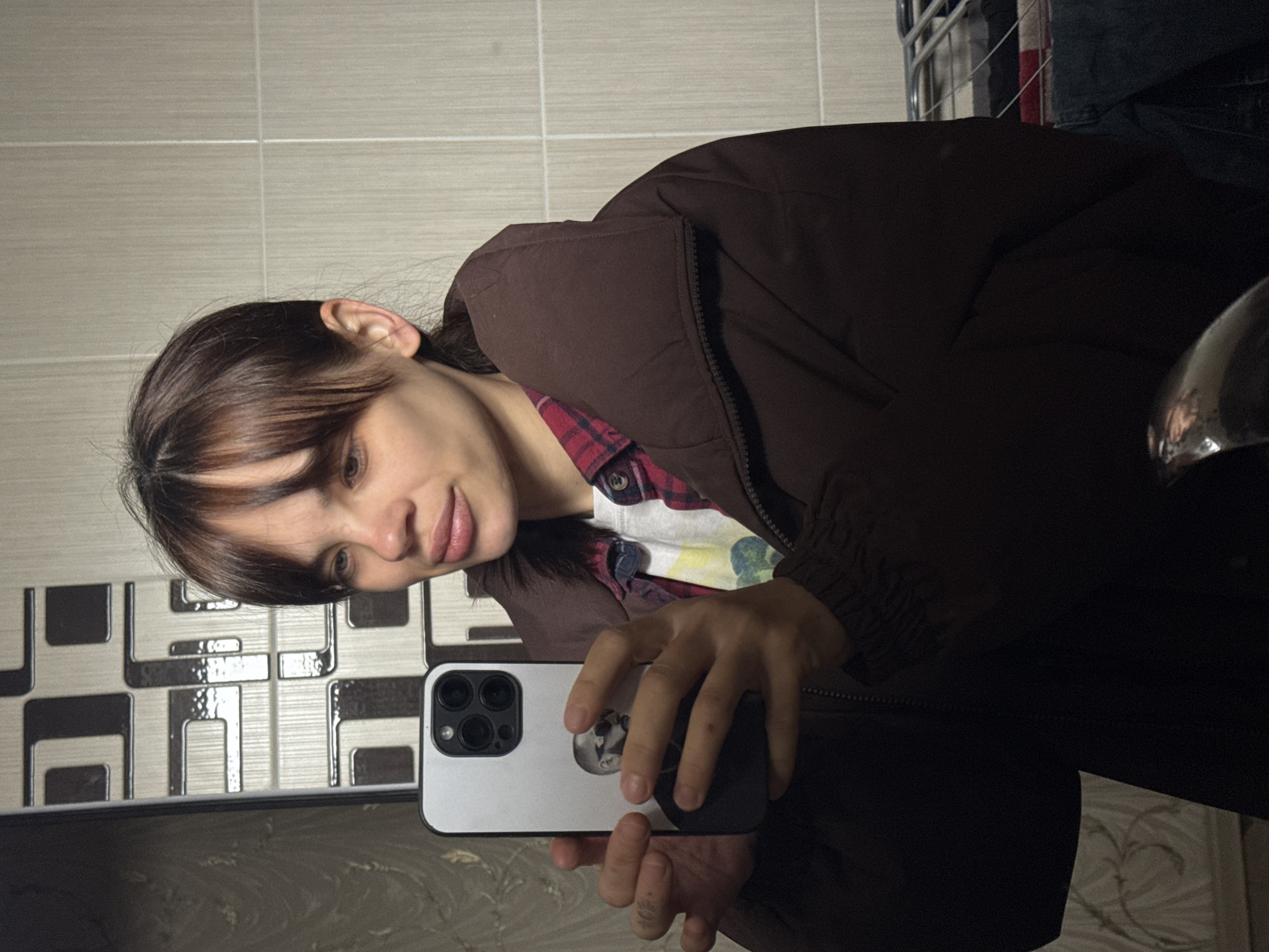 A woman is taking a selfie in a bathroom. The bathroom features neutral-colored tiled walls with a dark geometric pattern near the mirror. The woman is wearing a brown jacket over a red and black flannel shirt. She holds a light-colored smartphone with multiple rear cameras. The background hints at a cluttered interior with some metallic elements. The location is not explicitly identifiable as Rostov-on-Don, Russia, from the given visual information.