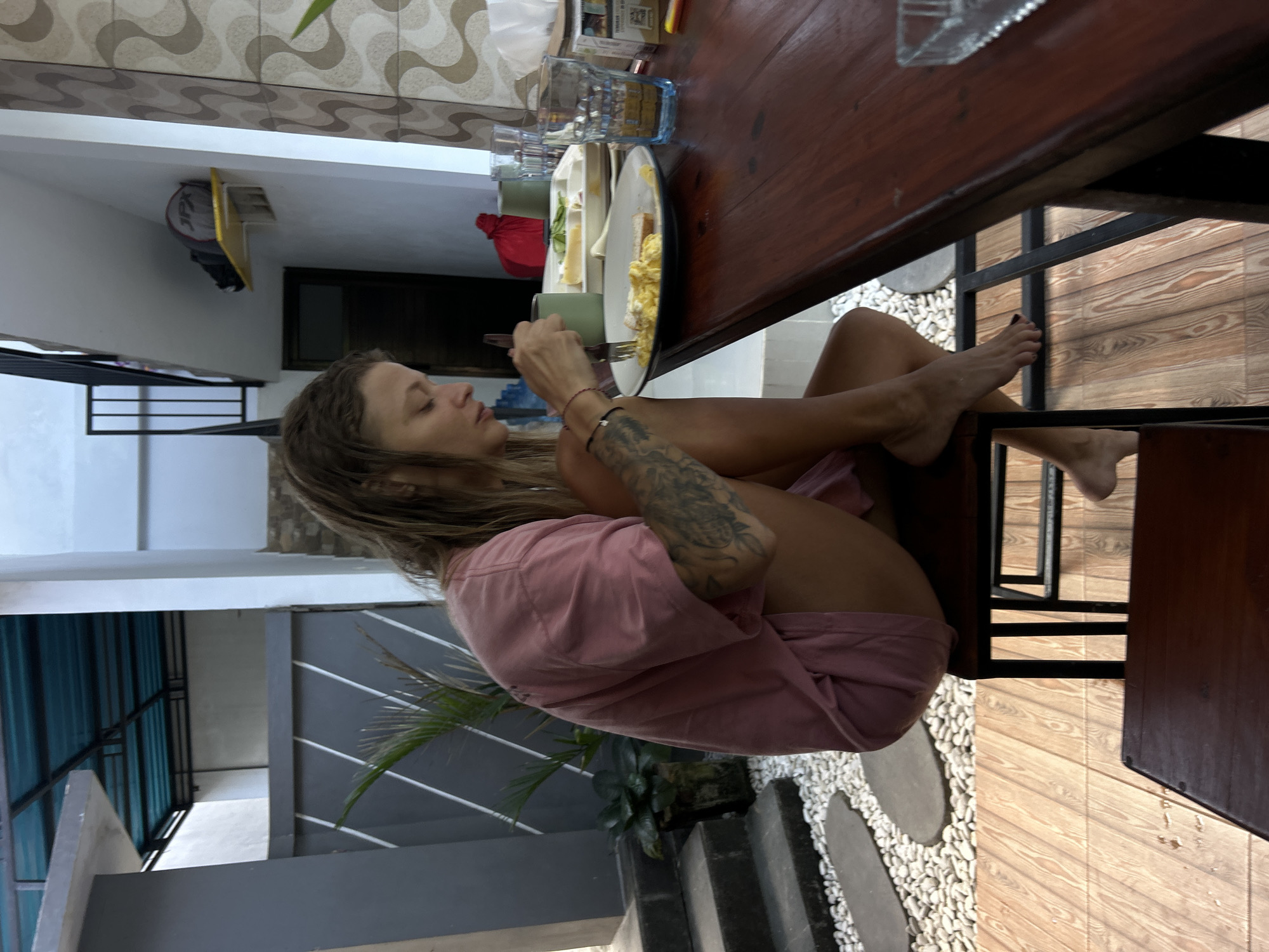 A woman with blonde hair and tattoos on her arm is sitting at a wooden table outdoors, eating scrambled eggs. She is wearing a pink t-shirt and shorts. The table has a plate of food, two glasses of water, and a package of cigarettes on it. The background shows a wall with a patterned design and a staircase leading up to a covered area. The ground is covered in white pebbles and wooden planks. The scene appears to be in North Kuta, Indonesia, given the architecture and plant life.