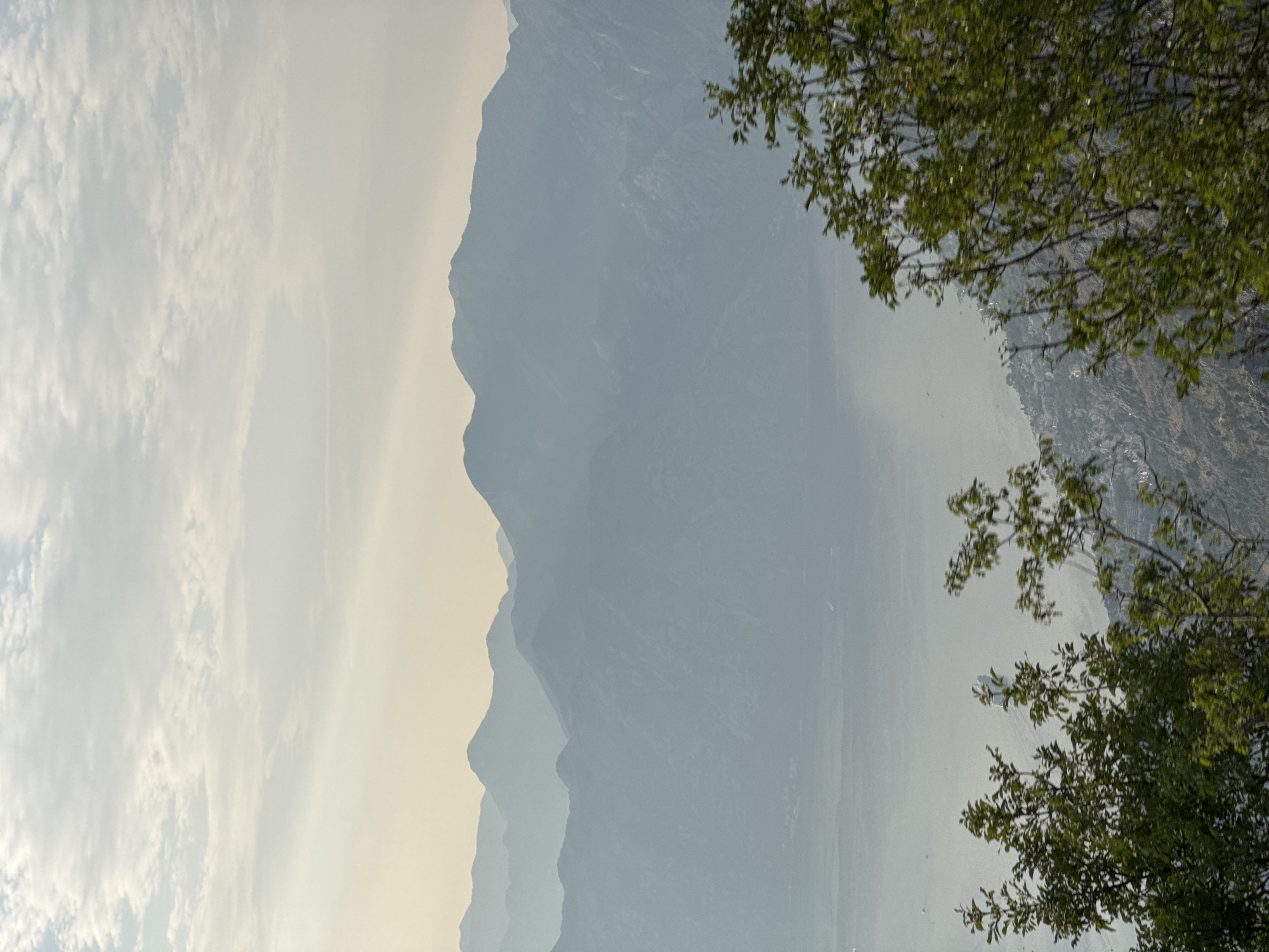 A vertical shot captures a hazy mountainous landscape from a high vantage point. The image is dominated by layers of muted blue mountains receding into the distance, creating a sense of depth. The sky above is a pale yellow-white with wispy clouds, suggesting either dawn or dusk. In the foreground, on the right and bottom sides, are the tops of lush green trees, their leaves slightly blurred, indicating movement or shallow depth of field. The foliage contrasts with the distant, soft-focused mountains. The lower part of the image shows a hint of a settlement or town nestled on a hillside, partially obscured by the haze and distance. The overall atmosphere is serene and natural, with a focus on the grand scale of the natural environment. The location is identified as Žanjev Do, Montenegro.