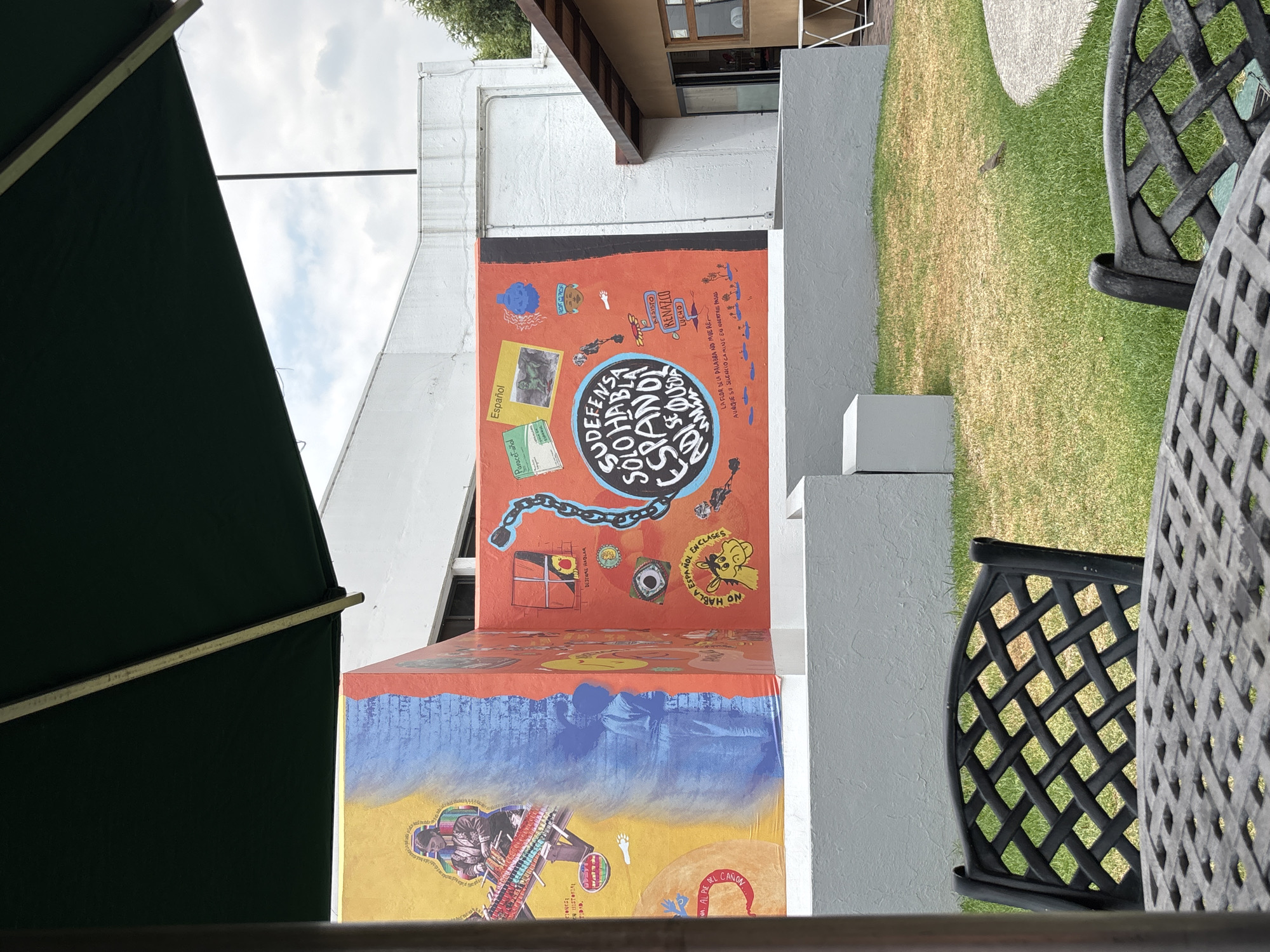 Here's a detailed description of the image:
**Location:** San Andrés Cholula, Mexico
**Scene:** The image captures a close-up view of an outdoor patio setting, prominently featuring a colorful mural on a building wall. There are black, decorative metal chairs and a table in the foreground, partially obscuring the view.
**Mural:** The mural spans two levels of the wall. The upper section has a red background with various painted elements, including written phrases, images of feet, chains, and cartoon-like faces. One prominent phrase within a blue circle reads, "DEFENSA SU SOLO HABLA ESPAÑOL ADIOS." Other written words in the mural include "EN HISTORIA TIDAD," "AL PIE DEL CAÑON," "DESENME HABLAR EN CLASES," "PIDLA ESPAÑOL," "Paracet-añol," "Español," "RENAZCO," and "AESISTO." The bottom section of the mural features a blue background transitioning into a yellow one, displaying a painted depiction of a person playing a musical instrument alongside other visual elements.
**Environment:** The patio is partially shaded by a dark green umbrella visible on the left side of the image. In the background, there is a glimpse of a lawn and other building structures. The buildings are painted white and are partially visible behind the mural.