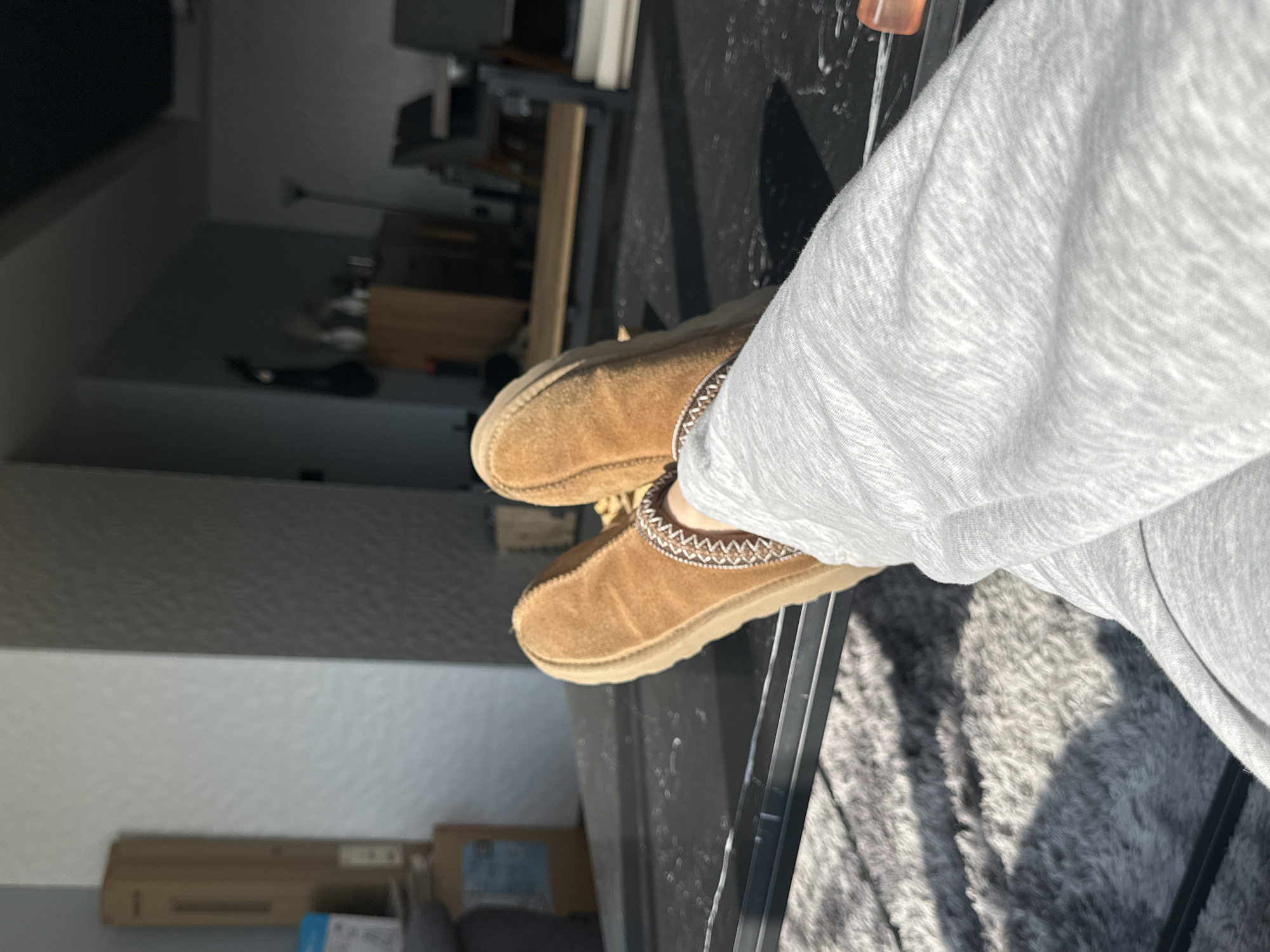 A close-up, slightly rotated, shot captures a person wearing light gray sweatpants and brown suede slippers with a decorative trim around the opening. They are positioned on a black surface, likely a table, with a gray, shaggy rug visible in the background. The environment includes a glimpse of a room with walls, and a structure that seems to be furniture, possibly a desk or storage unit. The location appears to be indoors, with natural light streaming in. There is no explicit evidence in the image to confirm the specific location as Huixquilucan de Degollado, Mexico.