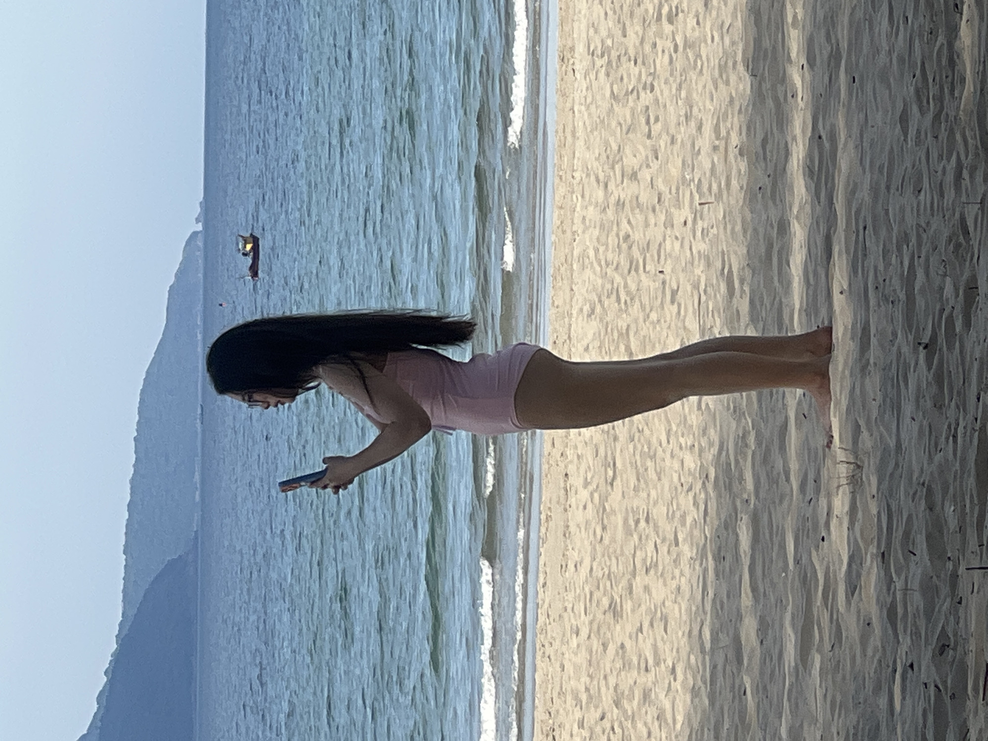 A young woman stands on a sandy beach, facing to her left, holding a smartphone in her hands. She is wearing a pink one-piece swimsuit and glasses. The ocean stretches out behind her, with gentle waves lapping at the shore. In the distance, mountains form a hazy backdrop against the sky, and a small boat is visible on the water. The beach is covered in light-colored sand with some scattered debris. The scene suggests a relaxed day at the coast, possibly in Đà Nẵng, Vietnam, known for its beautiful beaches and coastal scenery.