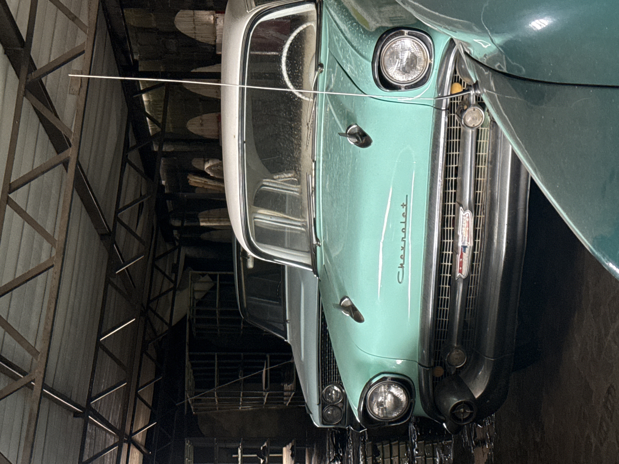 The image depicts the front of a vintage turquoise Chevrolet car parked indoors. The car is partially visible, showing the hood, grill, headlights, and windshield. The word "Chevrolet" is written in cursive on the hood. The background shows the interior of a building with metal beams and barrels. The location is unspecified, so I can't connect it to Kraaifontein, South Africa.
