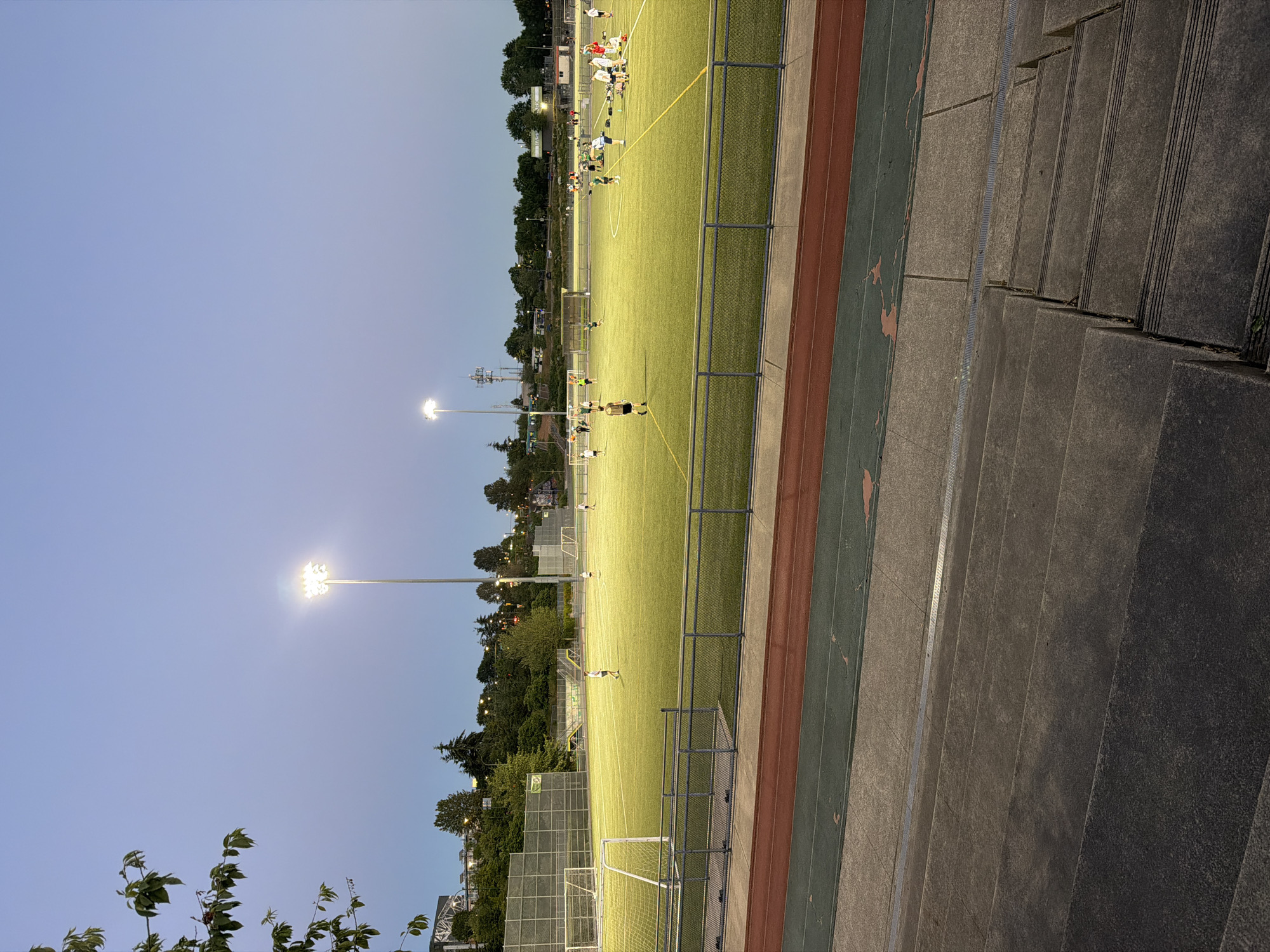 This image depicts a soccer field at dusk in Vancouver, Canada. The field is illuminated by tall stadium lights, casting a bright glow on the artificial turf. Several individuals, likely soccer players, are scattered across the field, some appearing to be engaged in practice or a casual game. A chain-link fence surrounds the field, and a running track with red and green lanes is visible in the foreground. Beyond the field, trees and urban structures are visible against the twilight sky. The perspective is from a slightly elevated position, suggesting the viewer is on a set of concrete bleachers or stairs.