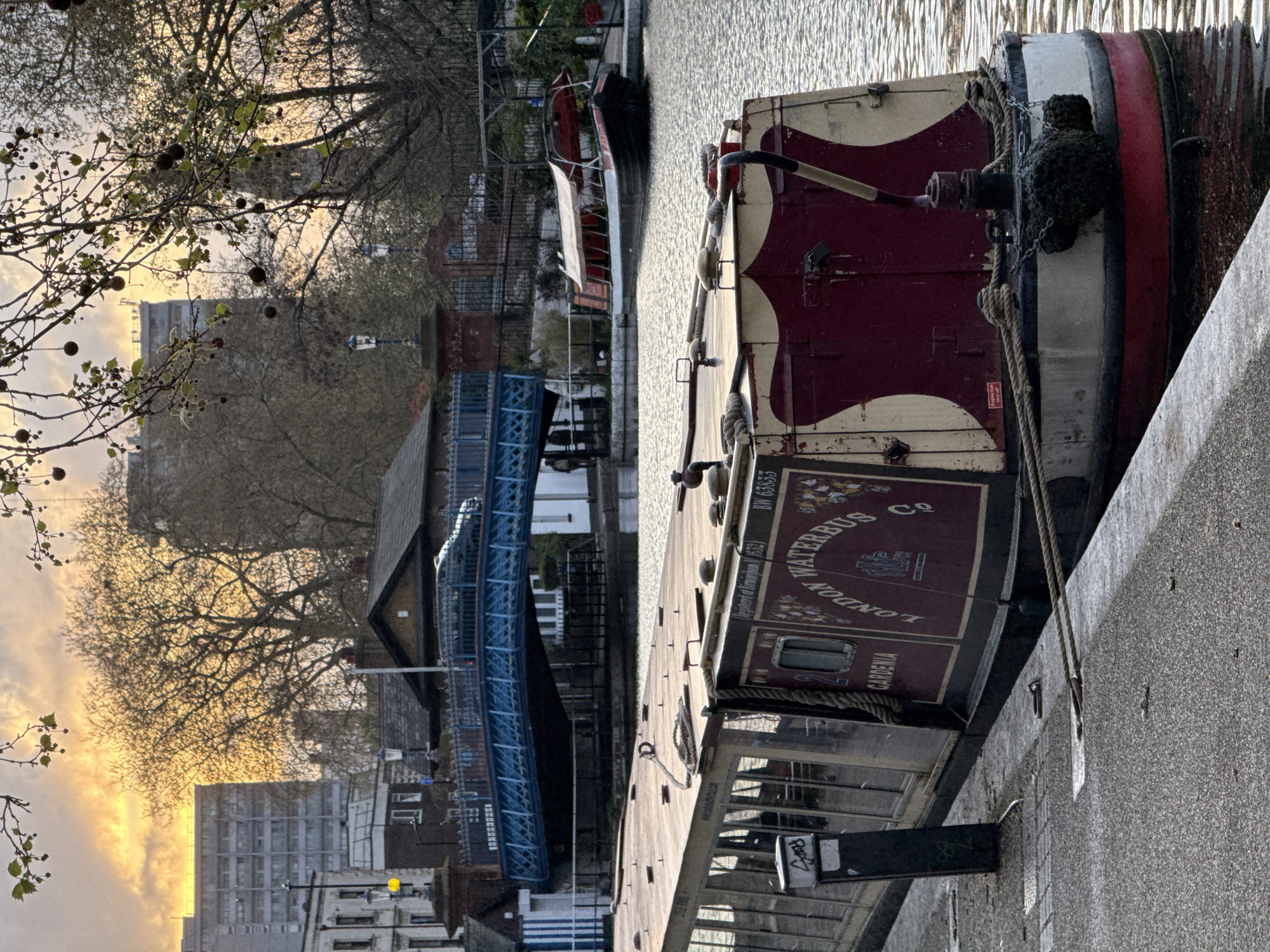 The image depicts a canal scene in London, United Kingdom, with a narrowboat ("Waterbus Co" - GARDENIA) docked alongside a paved walkway. The narrowboat has burgundy and cream colors, with a rope tied to a metal ring on the walkway. The canal water has a textured surface. In the background, a blue pedestrian bridge crosses the canal. Buildings are visible beyond the bridge, including high-rise apartments and older, brick structures. Trees with bare branches frame the scene. The sky is bright, suggesting either sunrise or sunset.