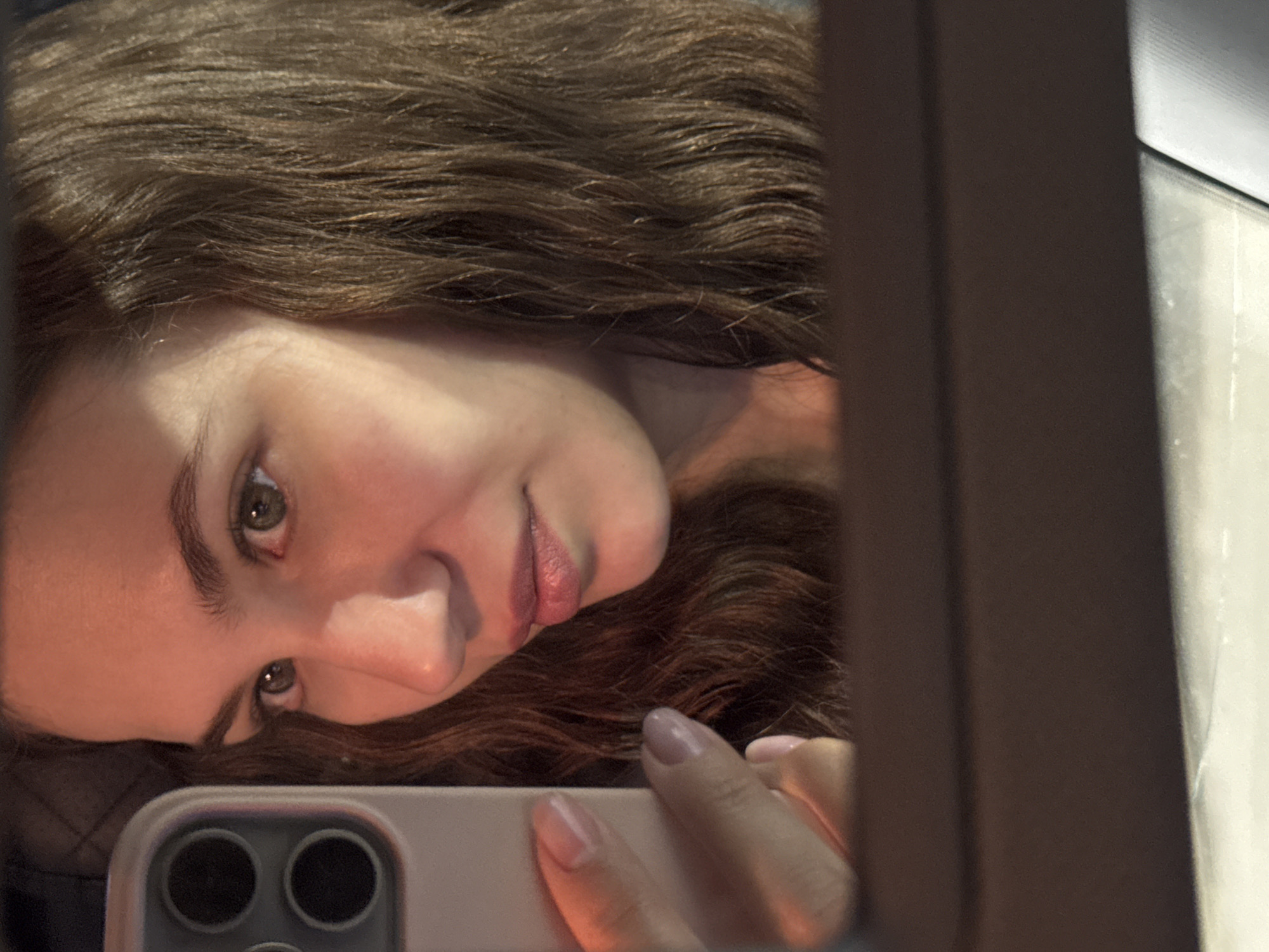 A young woman with long, wavy brown hair takes a selfie in what appears to be a car interior. She is holding a light-colored smartphone in her left hand, which is reflected in a mirror. The phone has a prominent camera module on the back. Her face is turned to the side, with her eyes looking into the mirror. The lighting in the image is warm and appears to be from artificial sources, casting a reddish hue on her skin. The background is blurred, suggesting a shallow depth of field. The lower portion of the image shows a dark surface, possibly the dashboard or a car seat, and a lighter, reflective surface which could be a car window. There is no indication of the specific location within Chaikovsky, Russia.