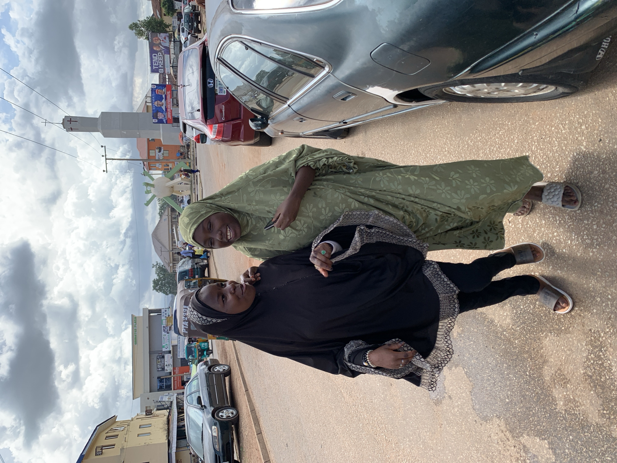 In this vertically oriented image, two young women are pictured on a street in Jos, Nigeria. The woman in the foreground is wearing a black hijab and abaya with a decorative, lacy trim on the sleeves and hem. She is looking to her left and smiling. The woman behind her is wearing a light green hijab and abaya patterned with small white flowers. She is also looking to her left and appears to be smiling. Both women are wearing sandals.
They are standing on a paved street next to a dark-colored car. Behind them, several other vehicles are visible, including a red car and a few auto-rickshaws. In the background, there are buildings and advertisements, including a large billboard that reads "TEND & KEEP" and mentions "WHO". Another sign reads "GAIRATES PLAZA". The sky is filled with dramatic clouds, suggesting it might be midday or late afternoon. The overall scene depicts a casual moment on a street in a busy urban area.