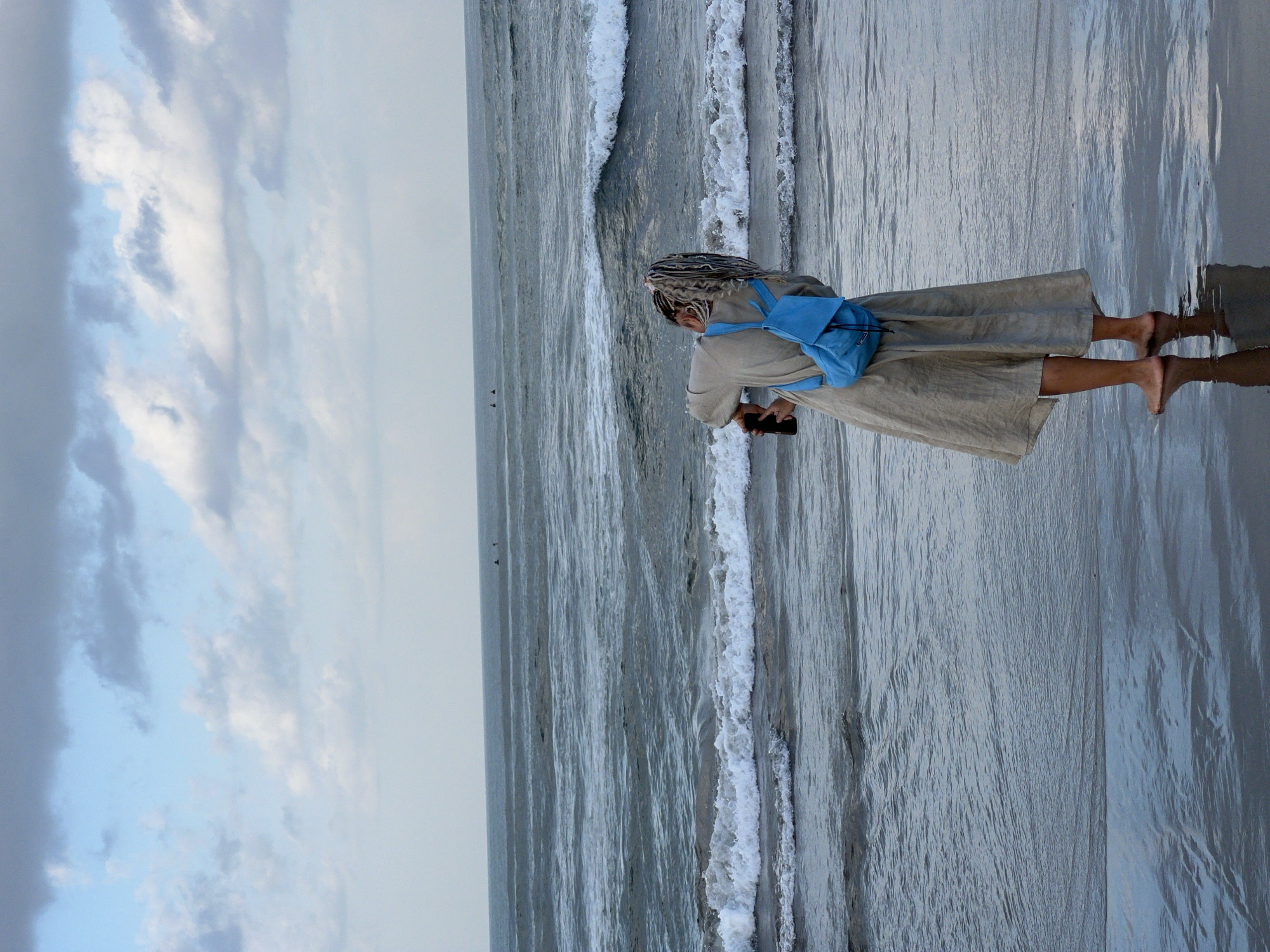 A person with light-colored dreadlocks stands on the wet sand of a beach, facing away from the camera. They are wearing a tan, loose-fitting dress and a bright blue backpack. The person is holding a dark-colored phone in their right hand, appearing to take a photograph of the ocean. The ocean is a deep blue-grey with white-capped waves rolling towards the shore. The sky above is a mix of light blue and grey clouds. The wet sand reflects the sky and the water. In the distance, two small figures can be seen on surfboards in the ocean. The scene suggests a location in North Kuta, Indonesia, likely a beach during overcast conditions.