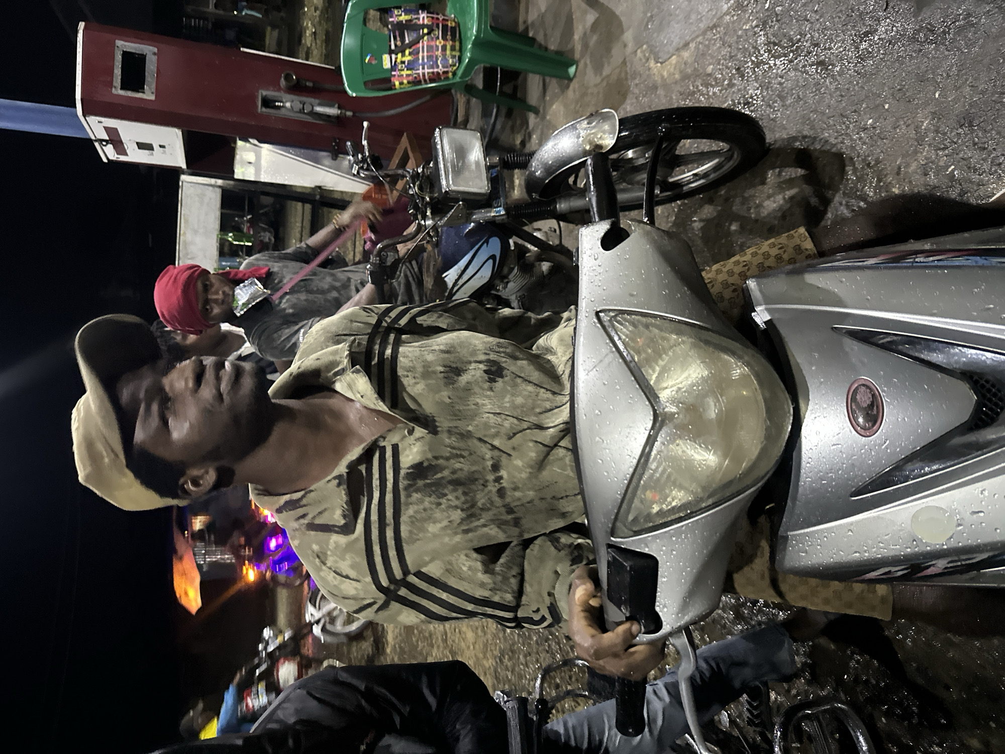 The image shows a man in Ikot Ekpene, Nigeria, wearing a tan cap and a camouflage jacket with black stripes, sitting on a silver scooter on a wet concrete surface. He's holding the scooter's handlebars. Another man wearing a red headscarf is nearby near a fuel pump. A green plastic chair and another motorcycle are in the background. Street lights illuminate the scene.