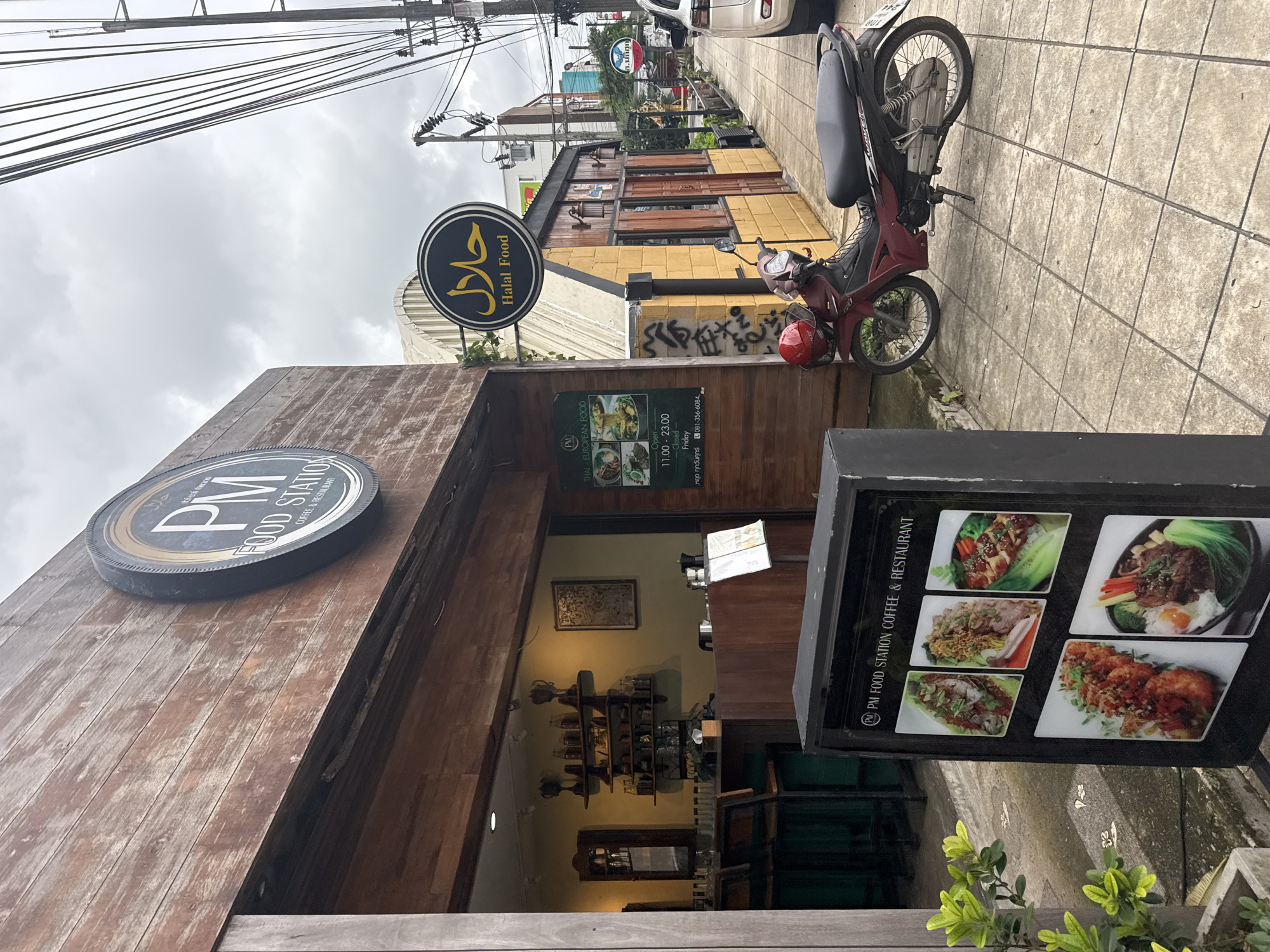 A roadside view in Chalong, Thailand, shows the exterior of "PM Food Station Coffee & Restaurant." The establishment features a wooden facade and a prominent circular sign with "PM FOOD STATION" and a coffee cup logo. Adjacent to it, a smaller yellow building displays a circular sign with Arabic script and "Halal Food." A red and black motorcycle is parked on the tiled sidewalk in front of the restaurant. A black A-frame sign near the entrance exhibits pictures of various food dishes. The sky is overcast, and utility wires are visible overhead.
