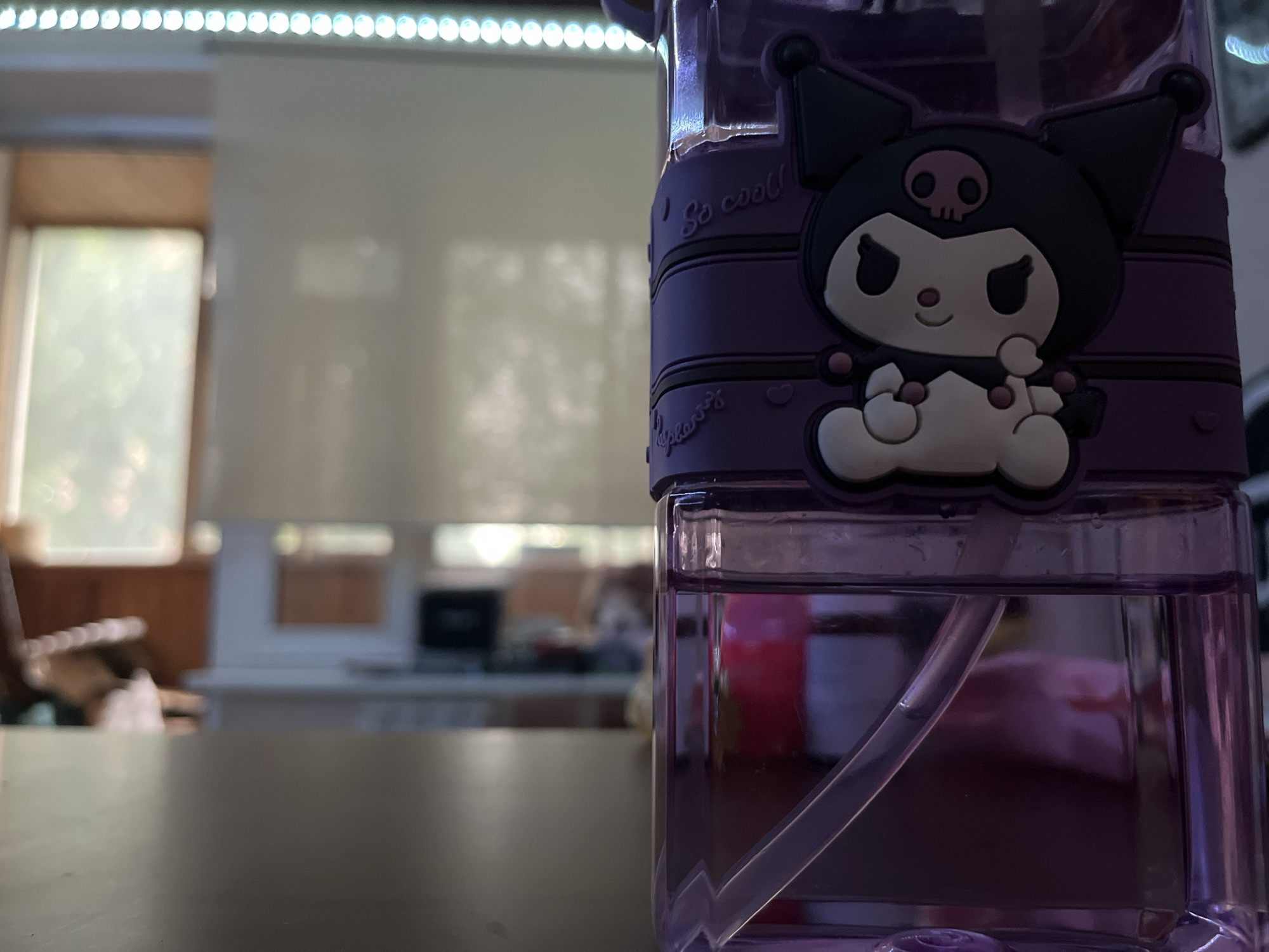 The image shows a close-up of a purple water bottle with a Kuromi character sticker on it. The bottle is filled with purple liquid and has a straw inside. The background is blurred and shows what appears to be a room with blinds on a window, a table, and some furniture. The lighting in the background suggests it might be daytime. The scene is indoors, in an indoor setting.