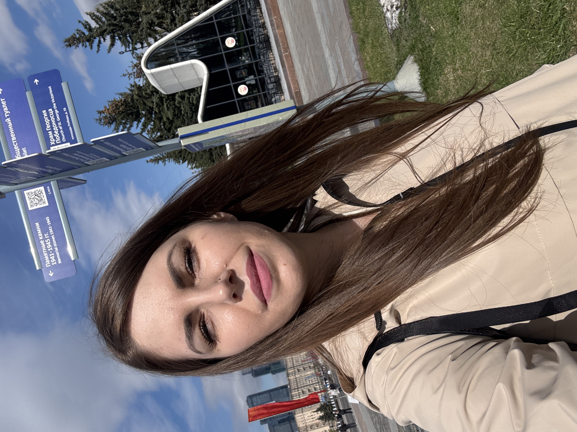 The image shows a woman posing for a selfie in the city of Moscow, Russia. The woman has long brown hair, fair skin, and is wearing a beige jacket with black trim. She is smiling slightly. In the background, there are directional signs pointing towards various locations, including the "Church of St. George the Victorious" and "Memorial Stones 1941-1945". Other buildings and a red flag can be seen in the distance. The sky is blue with some clouds.
