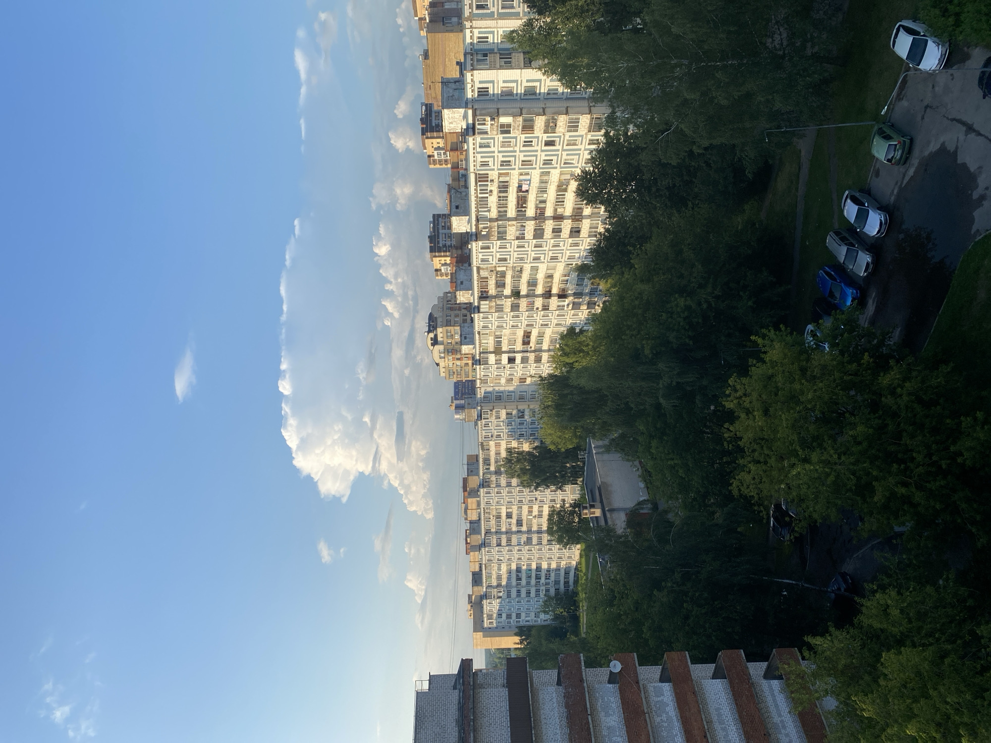 This is an aerial photograph of an urban residential area in Dubna, Russia. The scene is dominated by several multi-story apartment buildings, characteristic of Soviet-era architecture, with uniform facades featuring numerous windows and balconies. Lush green trees and foliage surround the buildings, forming a dense canopy that obscures parts of the lower levels. Several cars are parked on a paved area between the buildings and trees. The sky is a clear blue with scattered white cumulus clouds, suggesting fair weather. The lighting indicates it is likely late afternoon or early evening, with the sun casting long shadows and illuminating the buildings from the side.